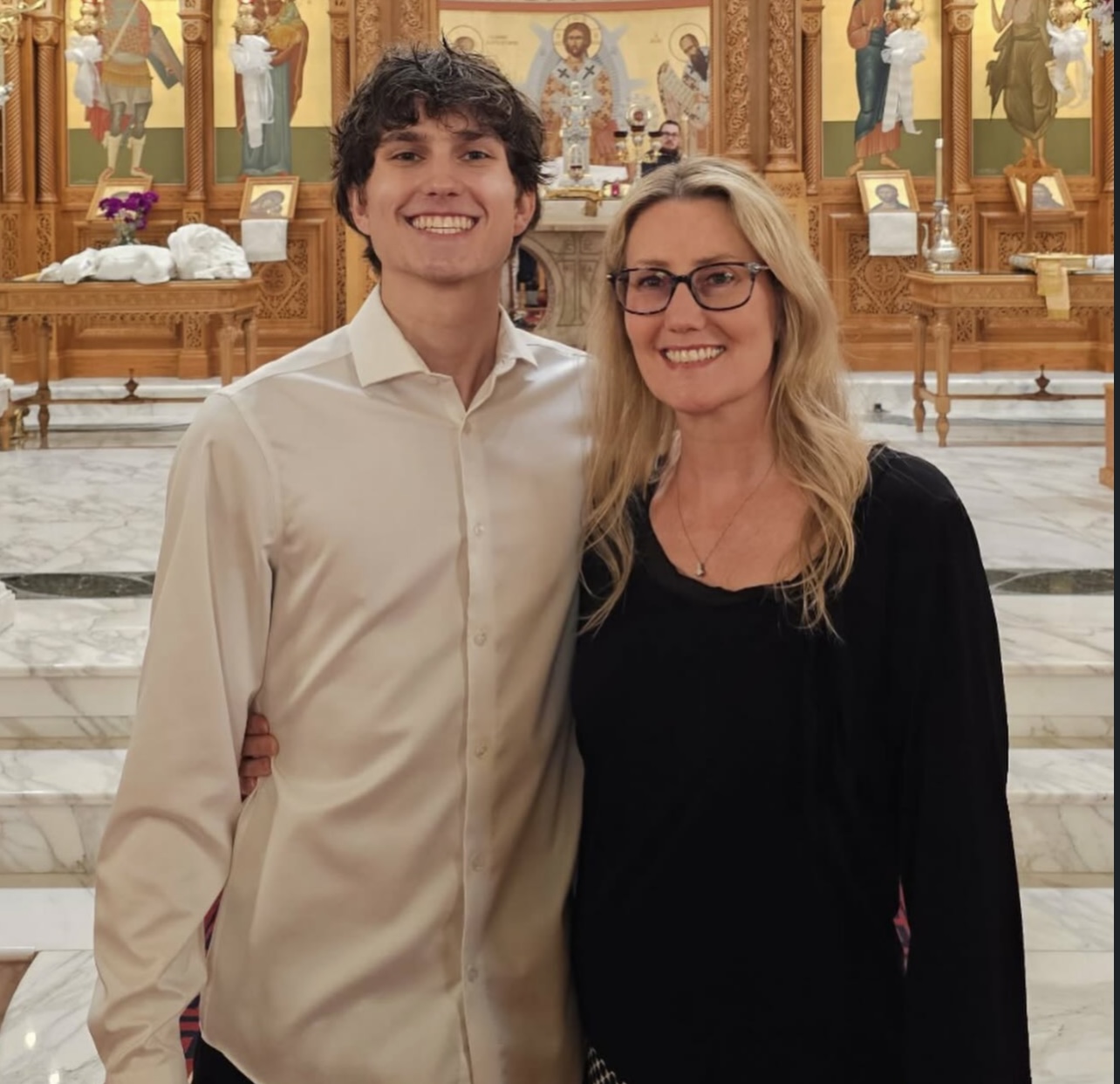 Two people smiling inside a church with religious icons and wooden decorations in the background.