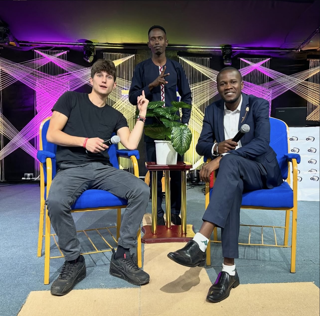 Three young men on stage with colorful lighting, two seated and one standing, holding microphones, in a modern setting.