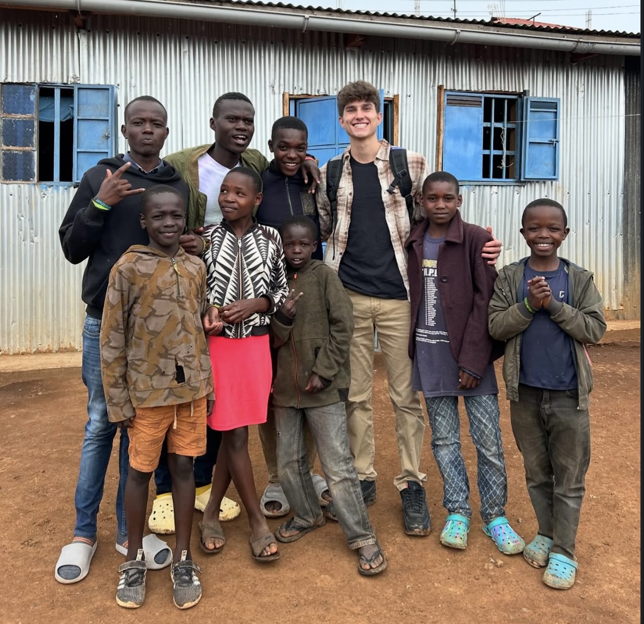 Group of ten children and one young adult standing outside in front of a building with blue windows, smiling and posing.