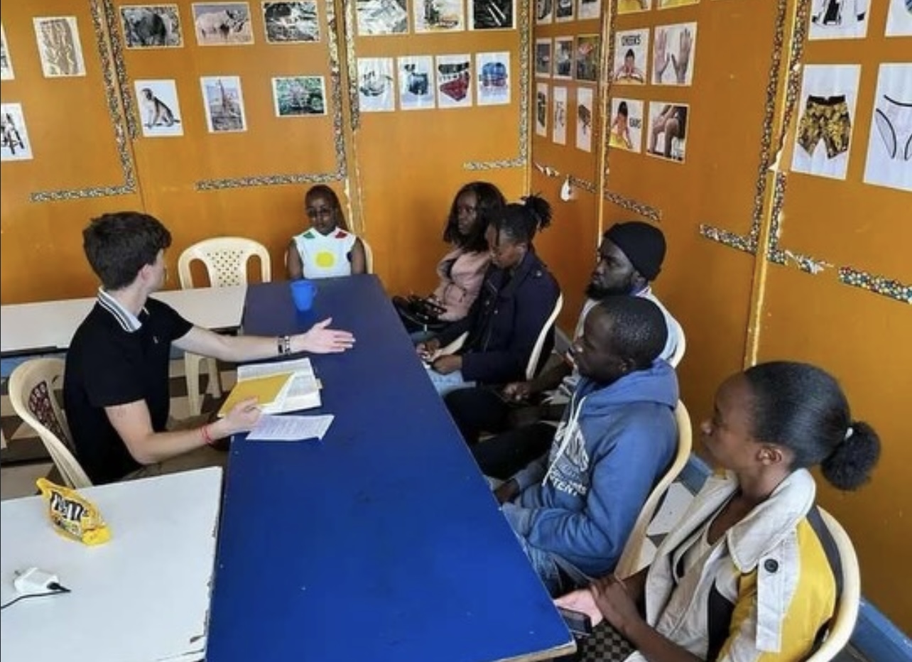 Group of children and a teacher sitting around a table in a classroom with yellow walls and artwork displays.