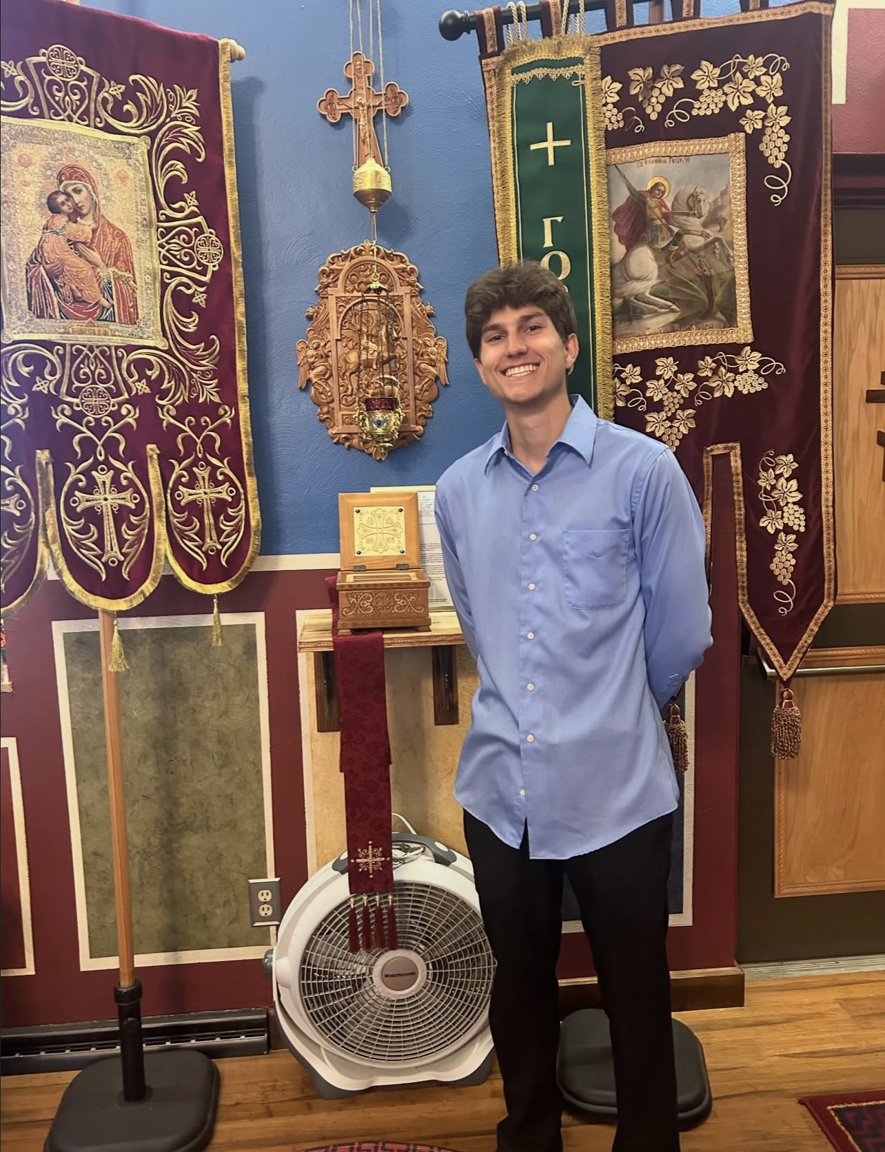 Young man in blue shirt smiling in a room with religious banners and icons, standing in front of a fan.