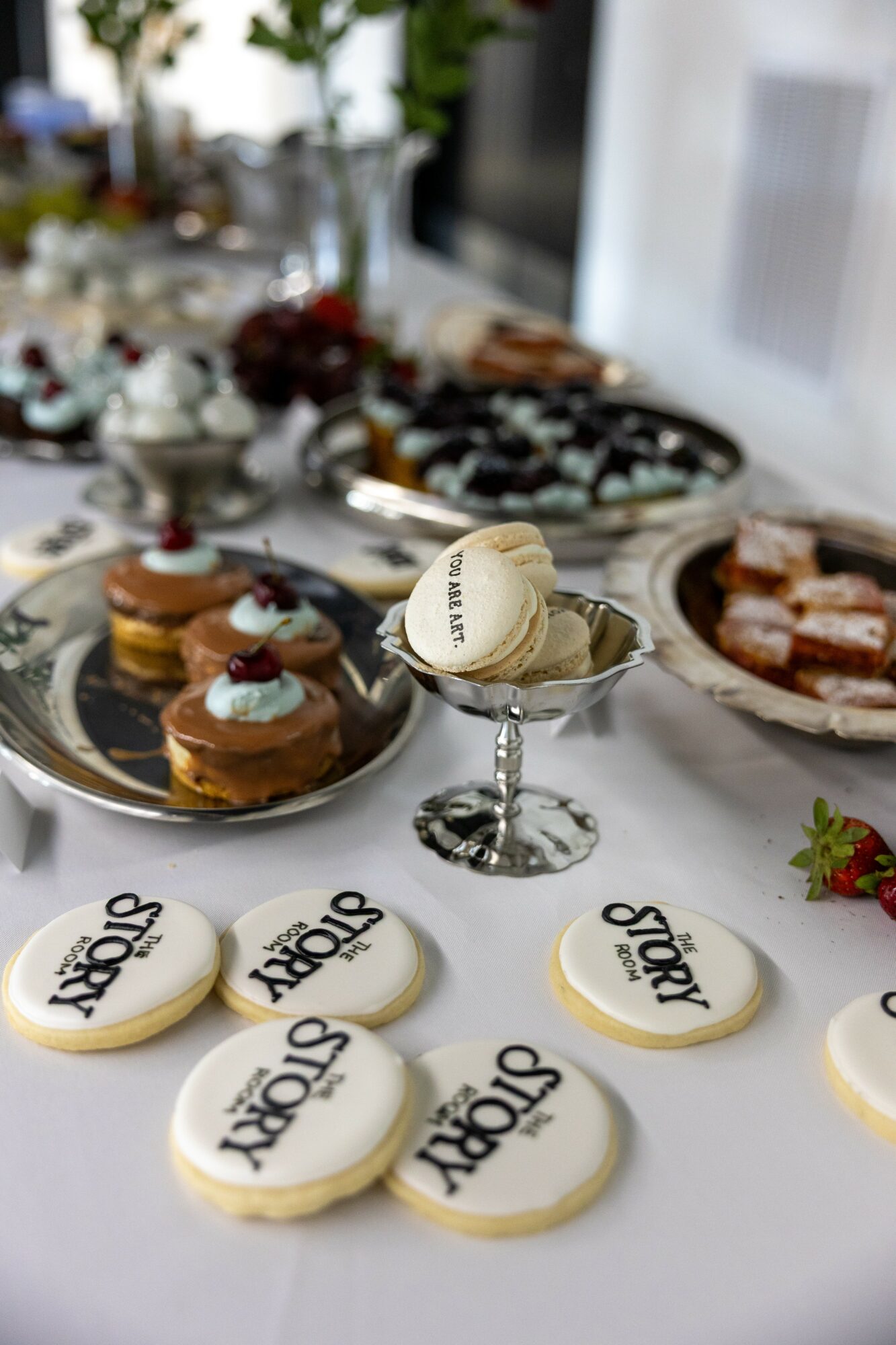 Dessert table with cookies, cupcakes, and other treats, with blurred background of a kitchen or dining area.