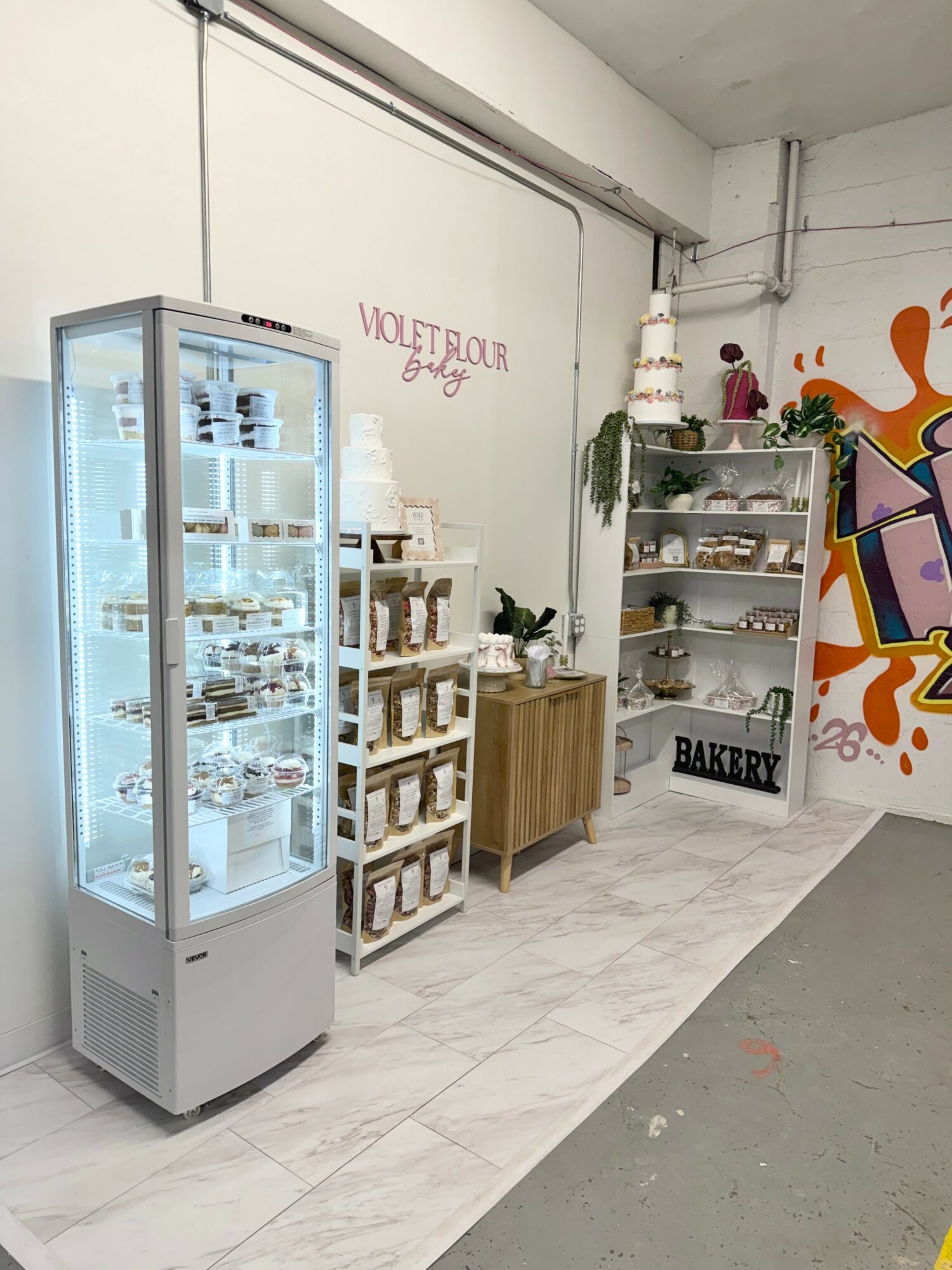 Display case with desserts, shelves with baked goods, and colorful wall art in a bakery setting.