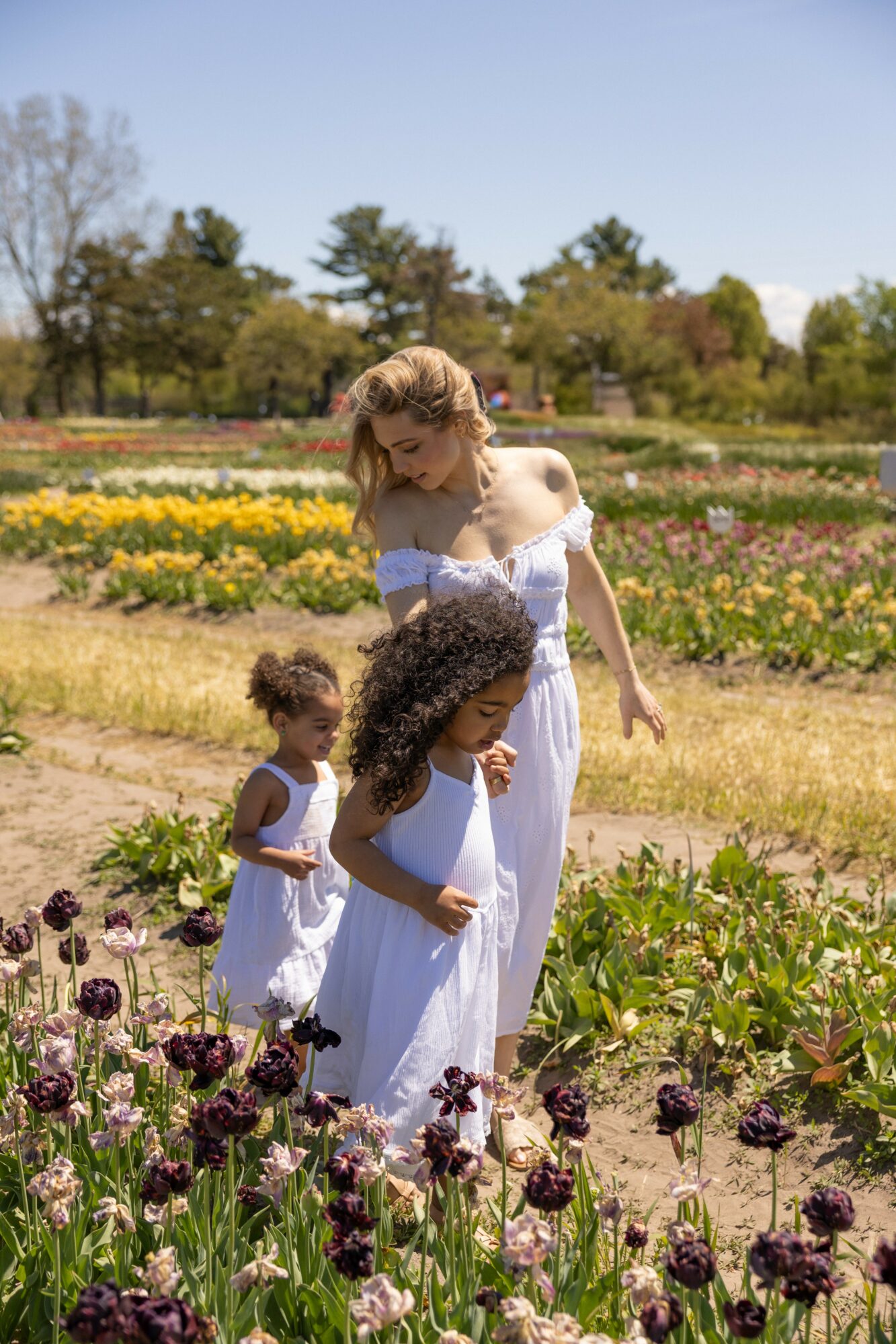 A woman and two children in white dresses walking through a colorful flower garden on a sunny day.