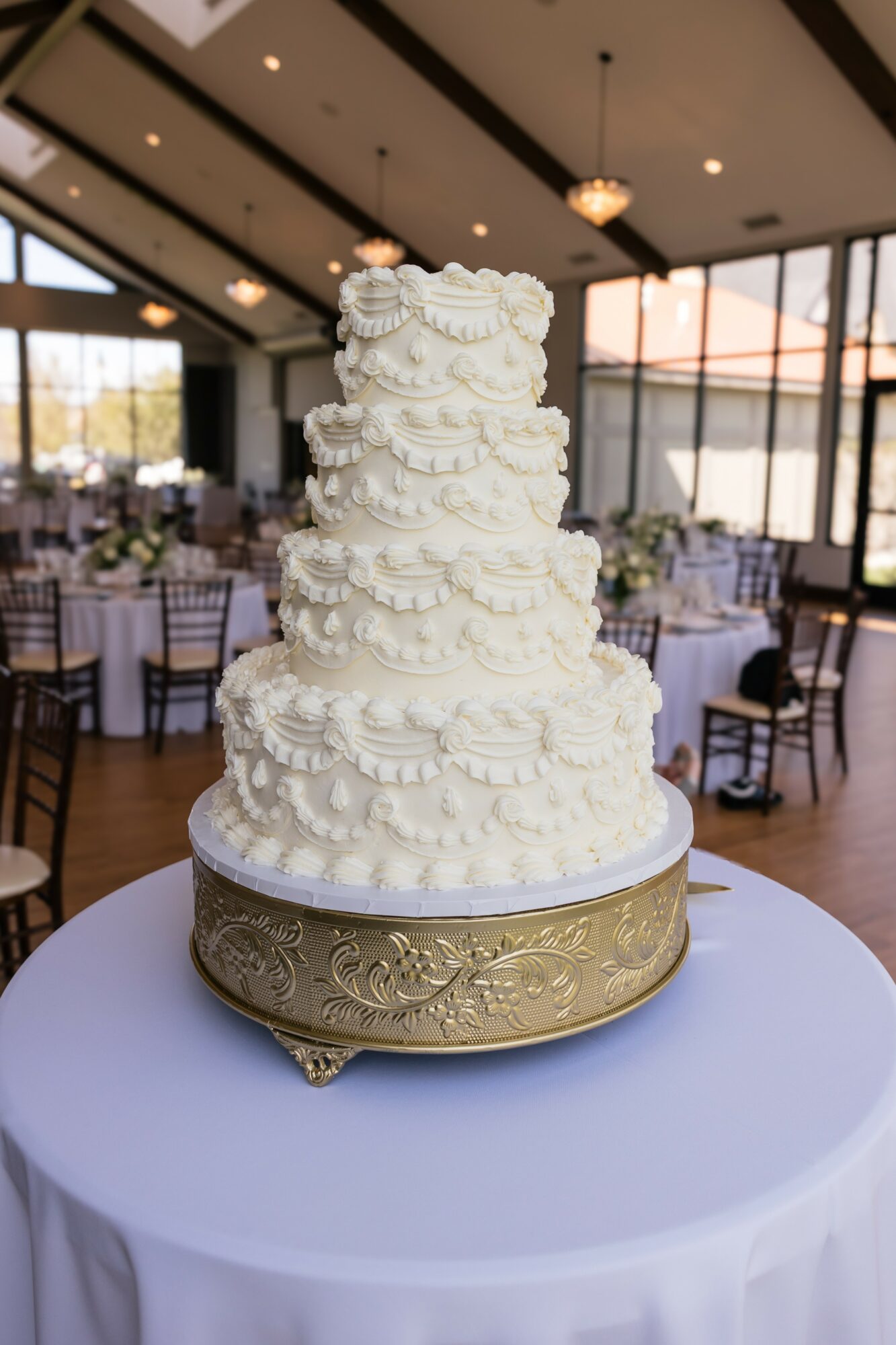 Tall, multi-tiered white wedding cake with decorative icing, on a round table in a decorated event hall.