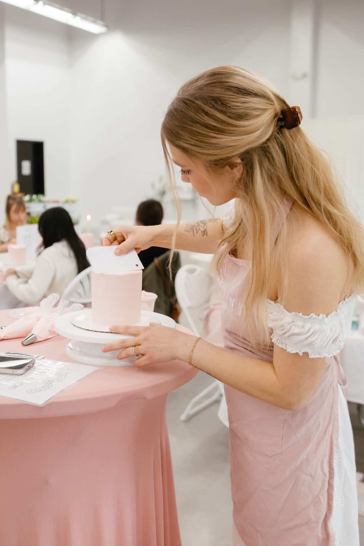 Woman in pink dress cutting a cake at a celebration, with people seated at a table in the background.