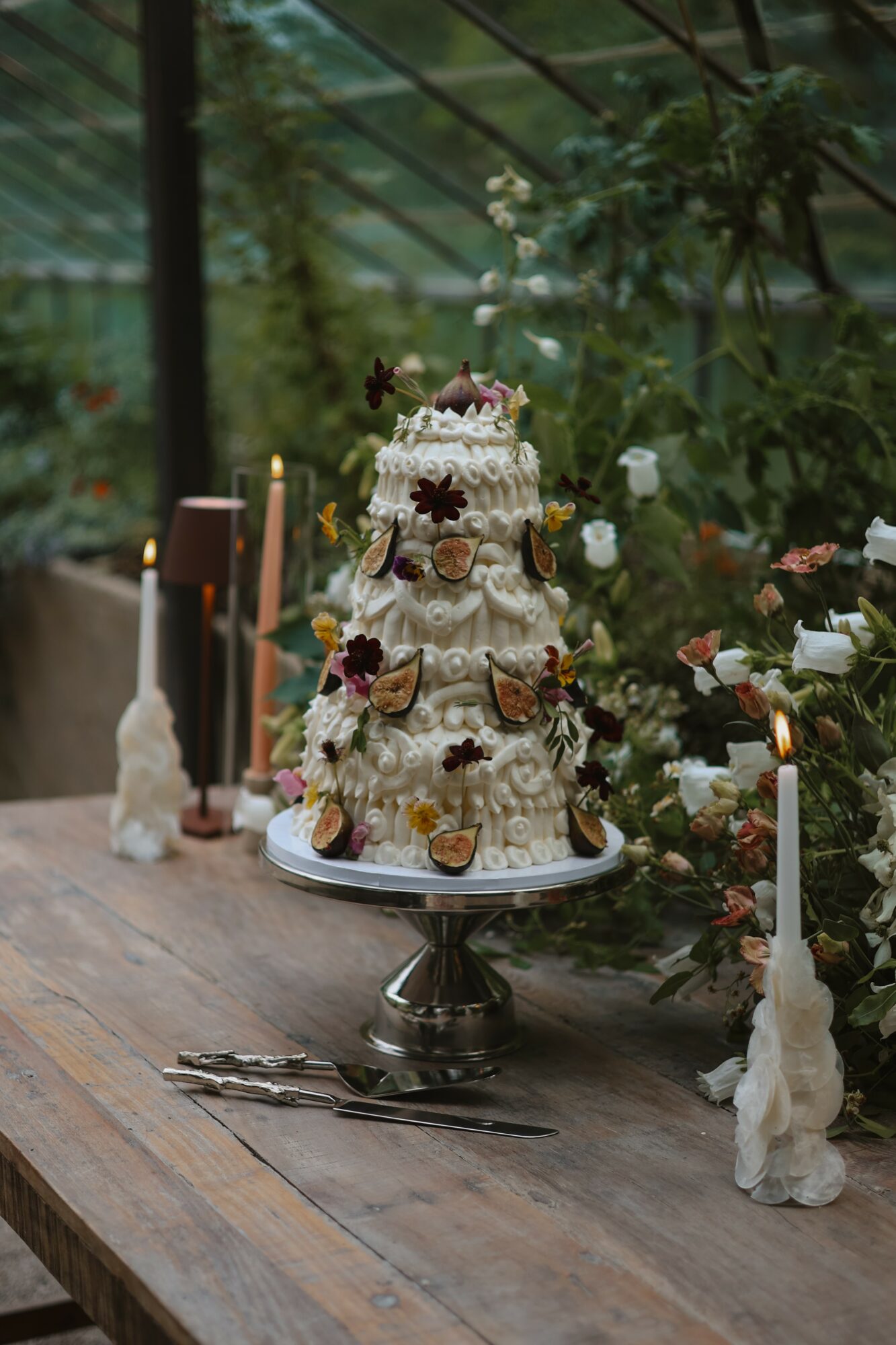 Three-tiered cake decorated with flowers and butterflies on a silver stand, surrounded by candles and flowers in a greenhouse.