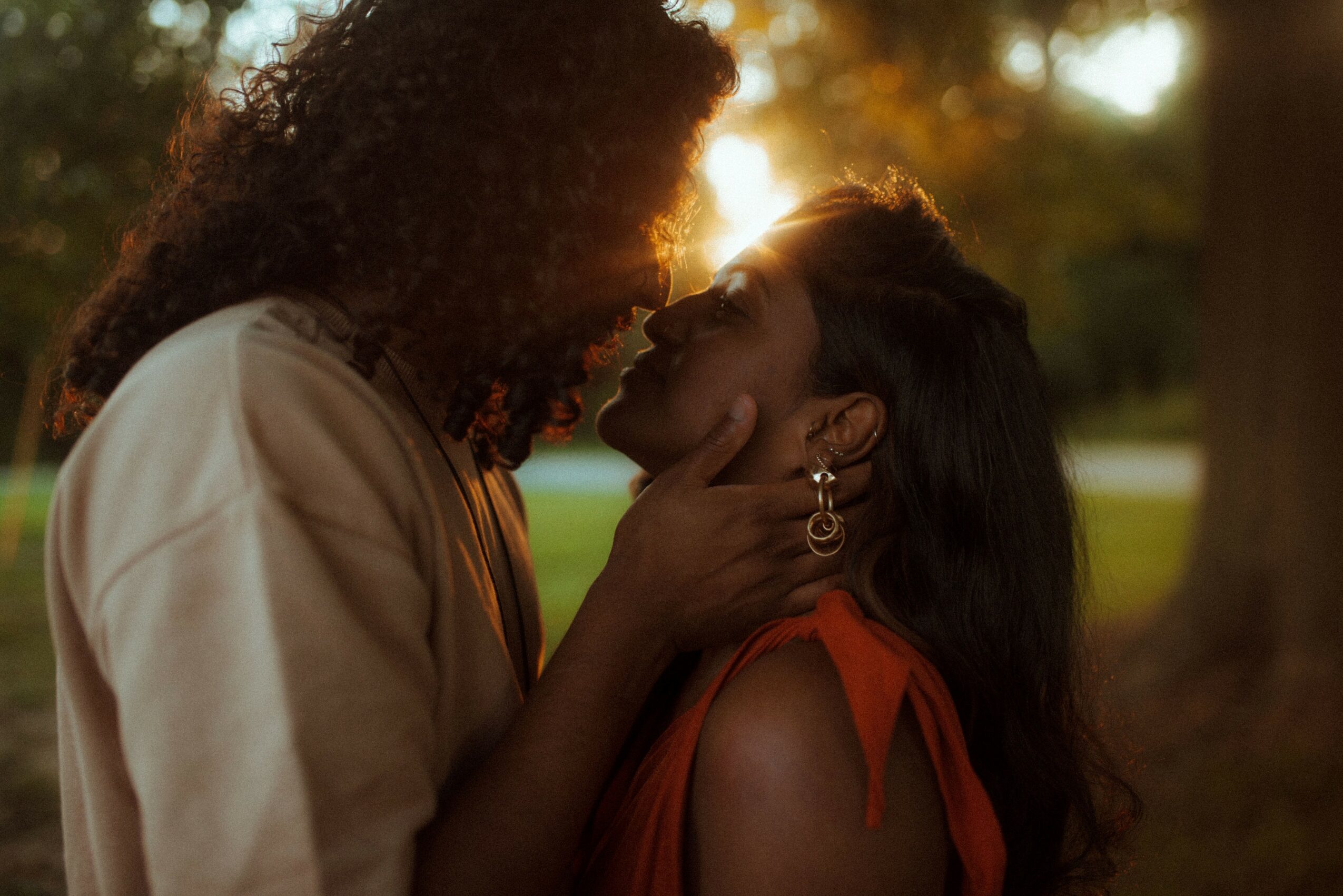 Two women face each other outdoors, one gently holding the other's face, with sunlight filtering through trees in the background.