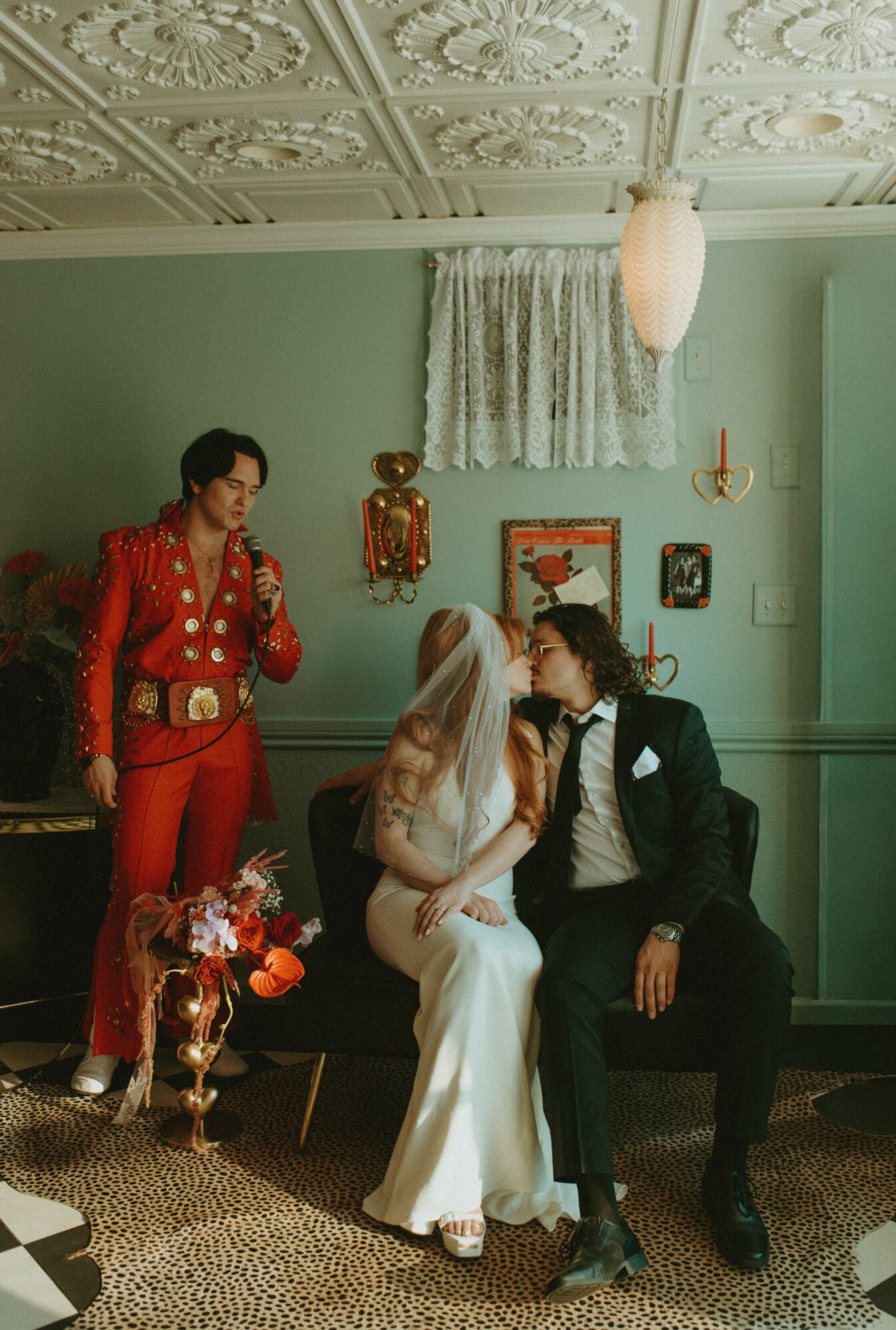 Three people in a room: one standing in a red outfit, two sitting on a bench, one in a white dress, the other in a black suit, with vintage decor.