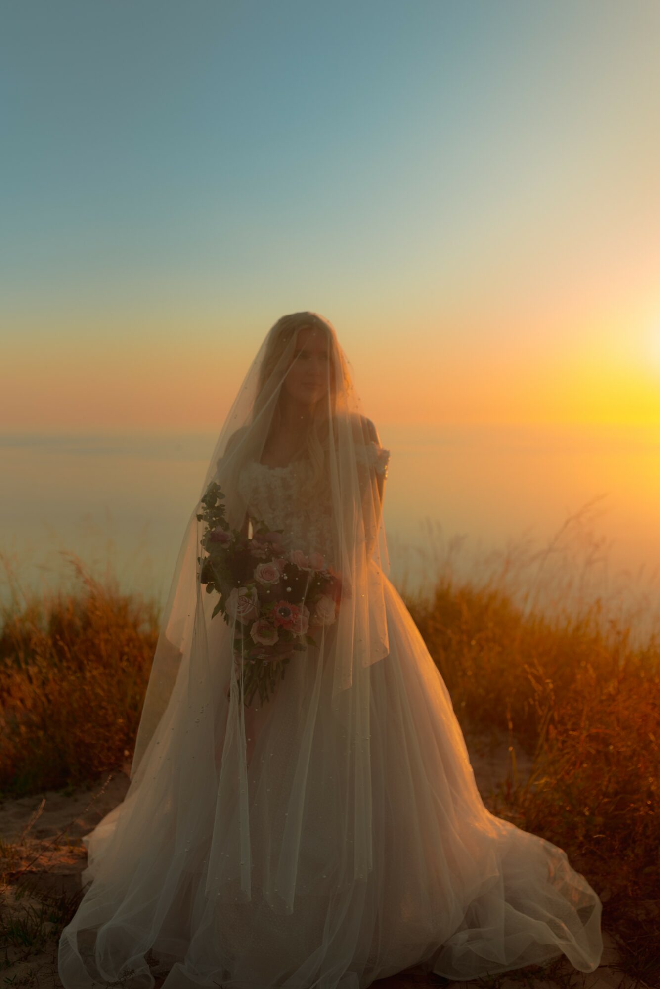 Bride in wedding dress holding a bouquet, standing outdoors at sunset with sky and landscape in background.