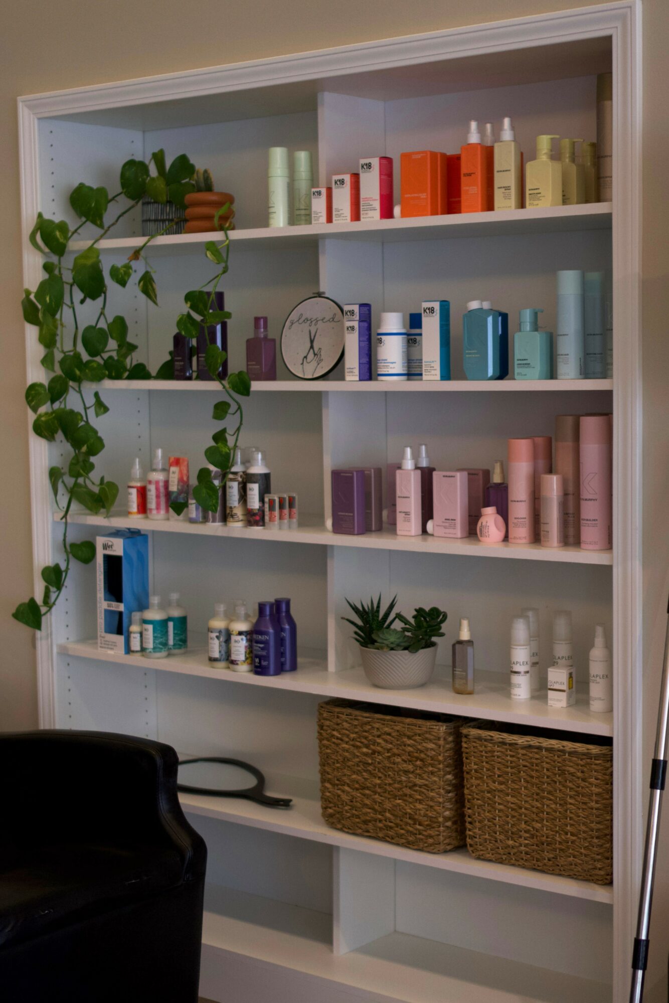 White bookshelf with colorful skincare products, a clock, potted plant, and two woven baskets, against a light-colored wall.