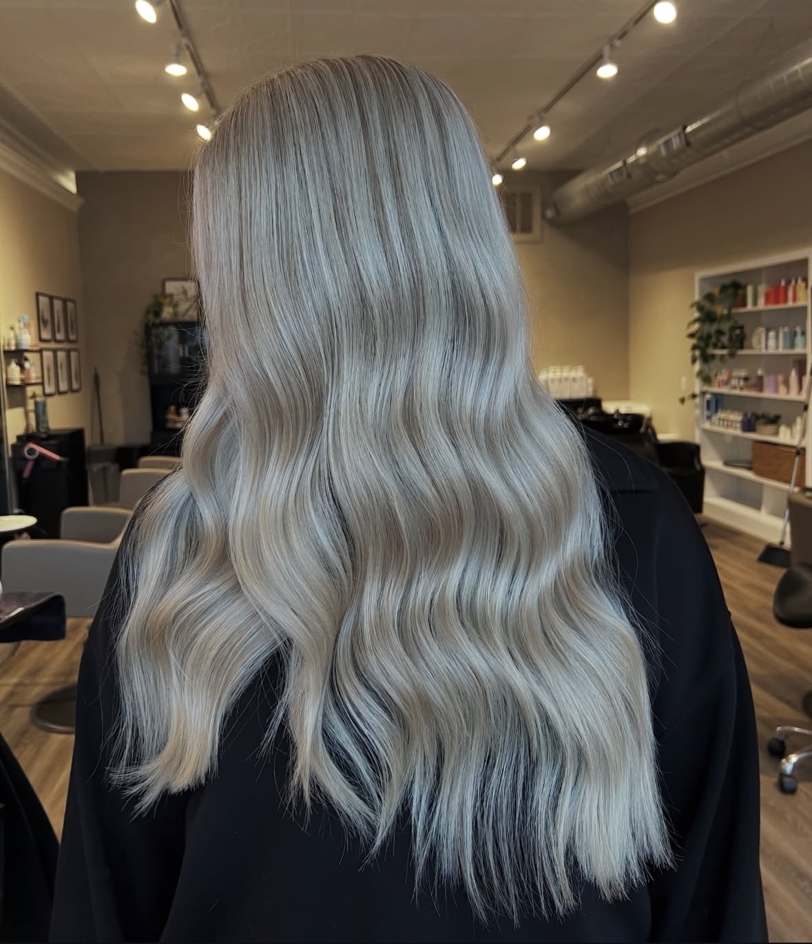 Back of a person with long, wavy, silver-blonde hair in a room with shelves and lighting.