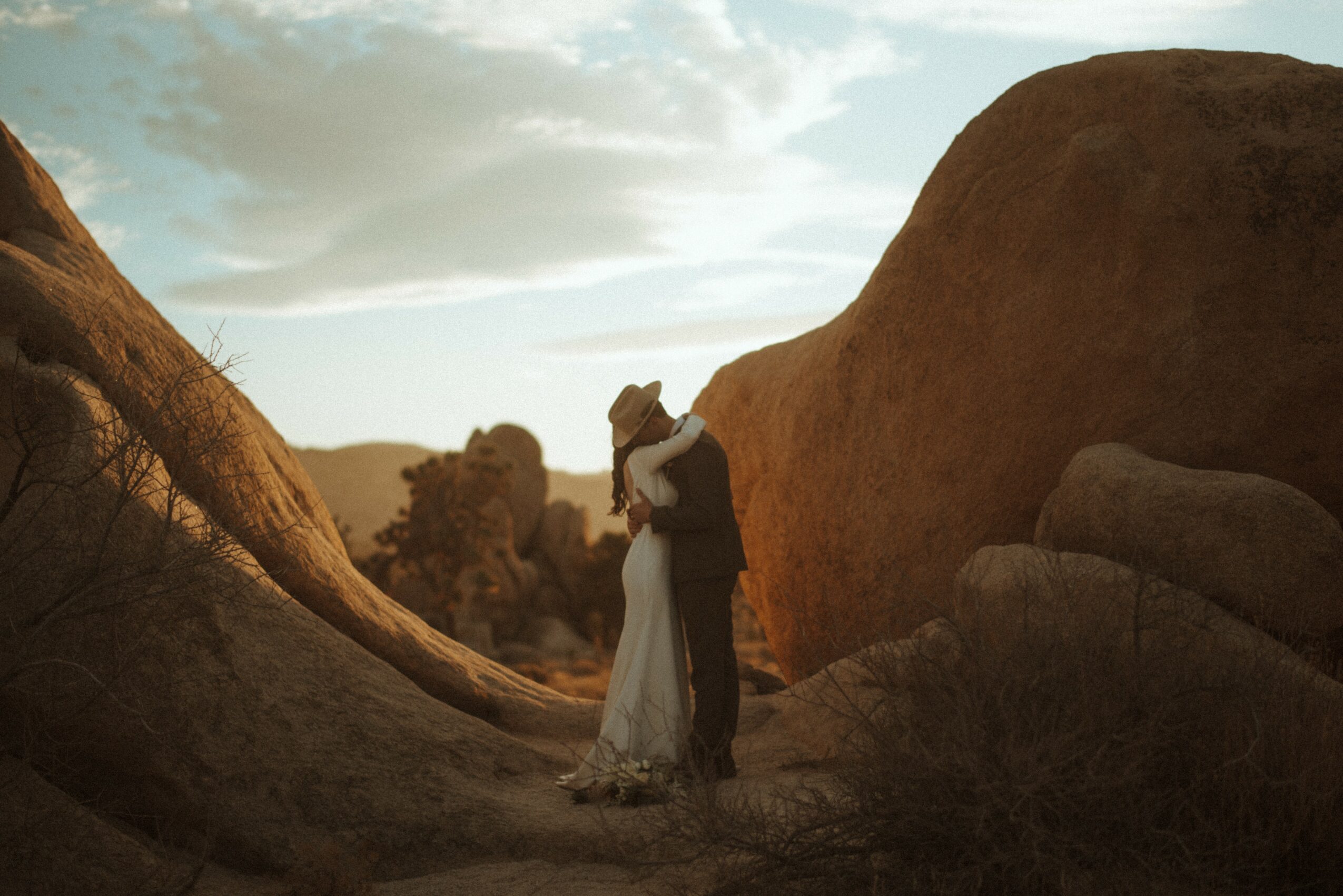 A couple stands close together between large rocks outdoors during sunset or sunrise.