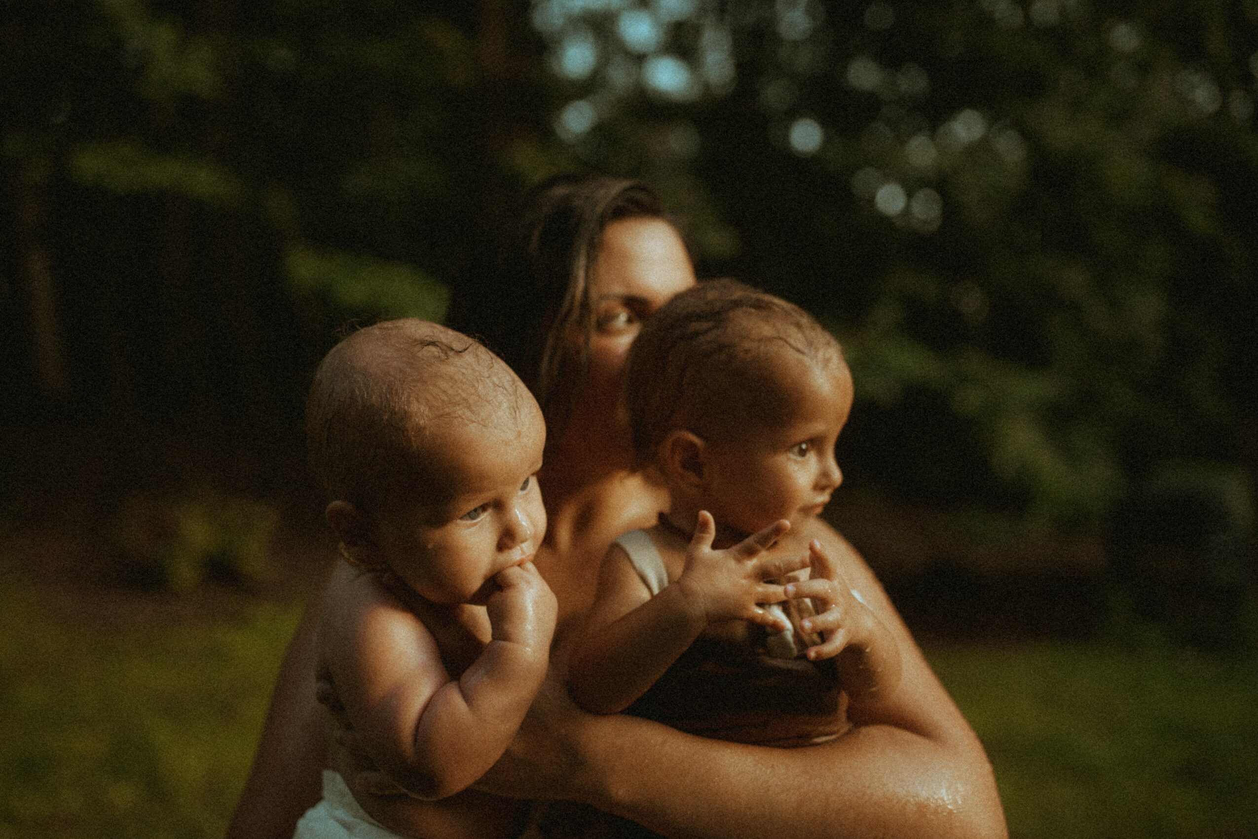 Woman holding two young children outdoors with trees in background, sunlight illuminating their faces.