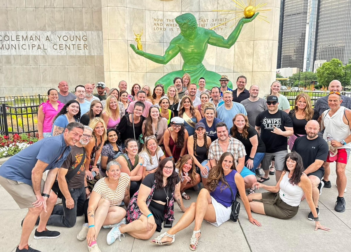 Group of people posing outdoors in front of a large green sculpture and a building, some smiling and making gestures.