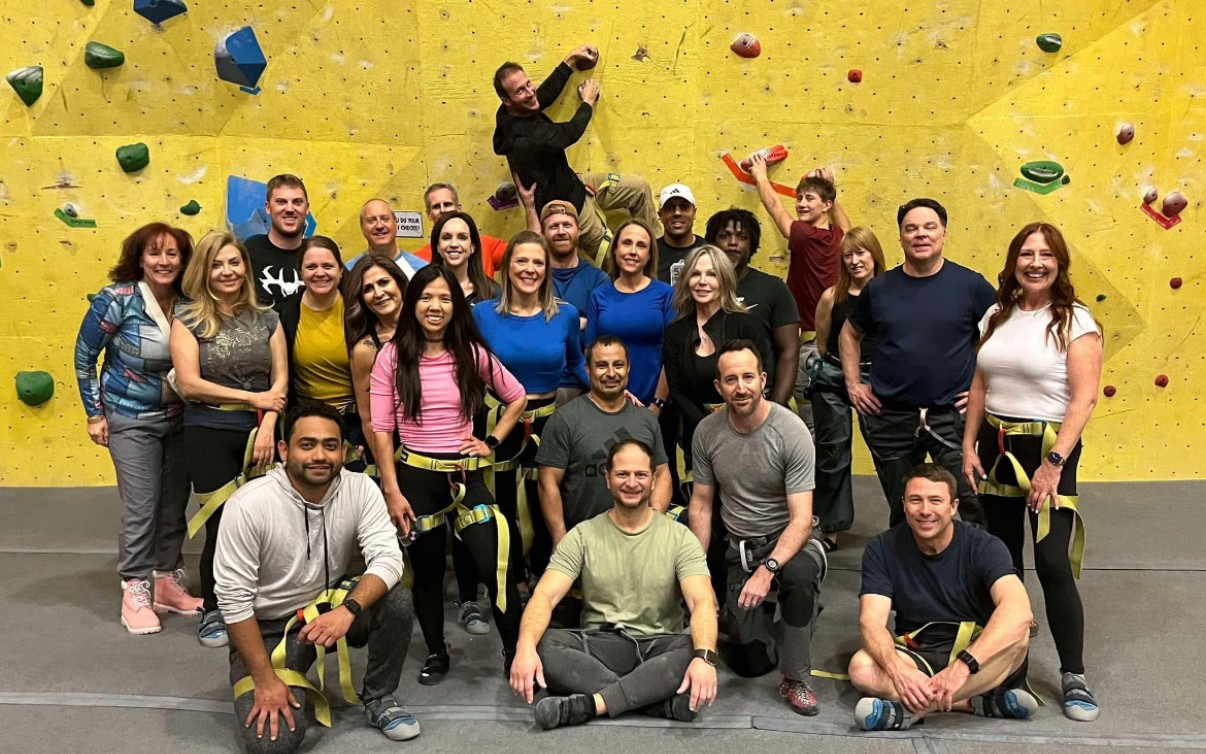 Group of people posing in front of an indoor climbing wall with colorful holds, some holding climbing gear, smiling.