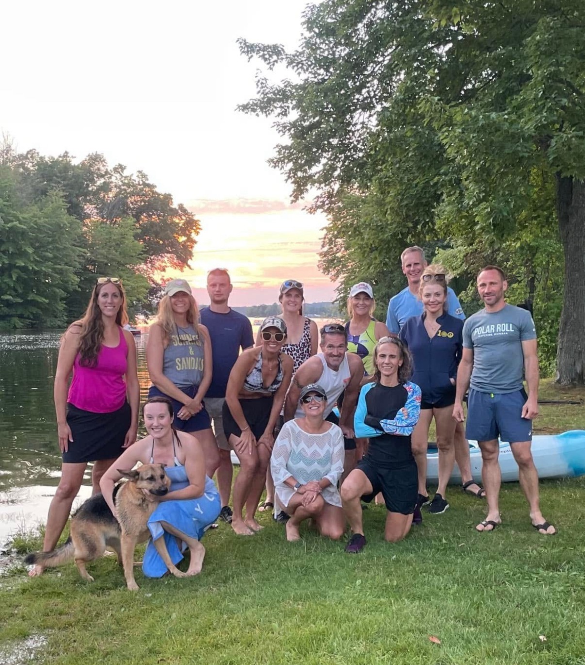Group of people outdoors near water, some sitting and some standing, with trees and a grassy area, during sunset.