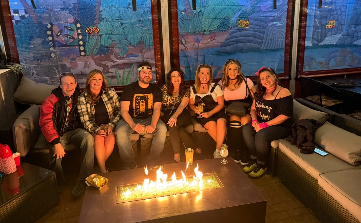 Group of seven people sitting on a couch in a room with colorful wall art, a fire feature, and a table with drinks.