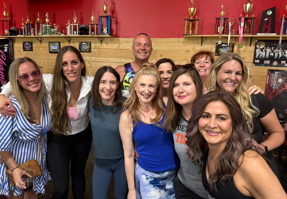 Group of ten smiling women and one man posing indoors with trophies on shelves behind them.