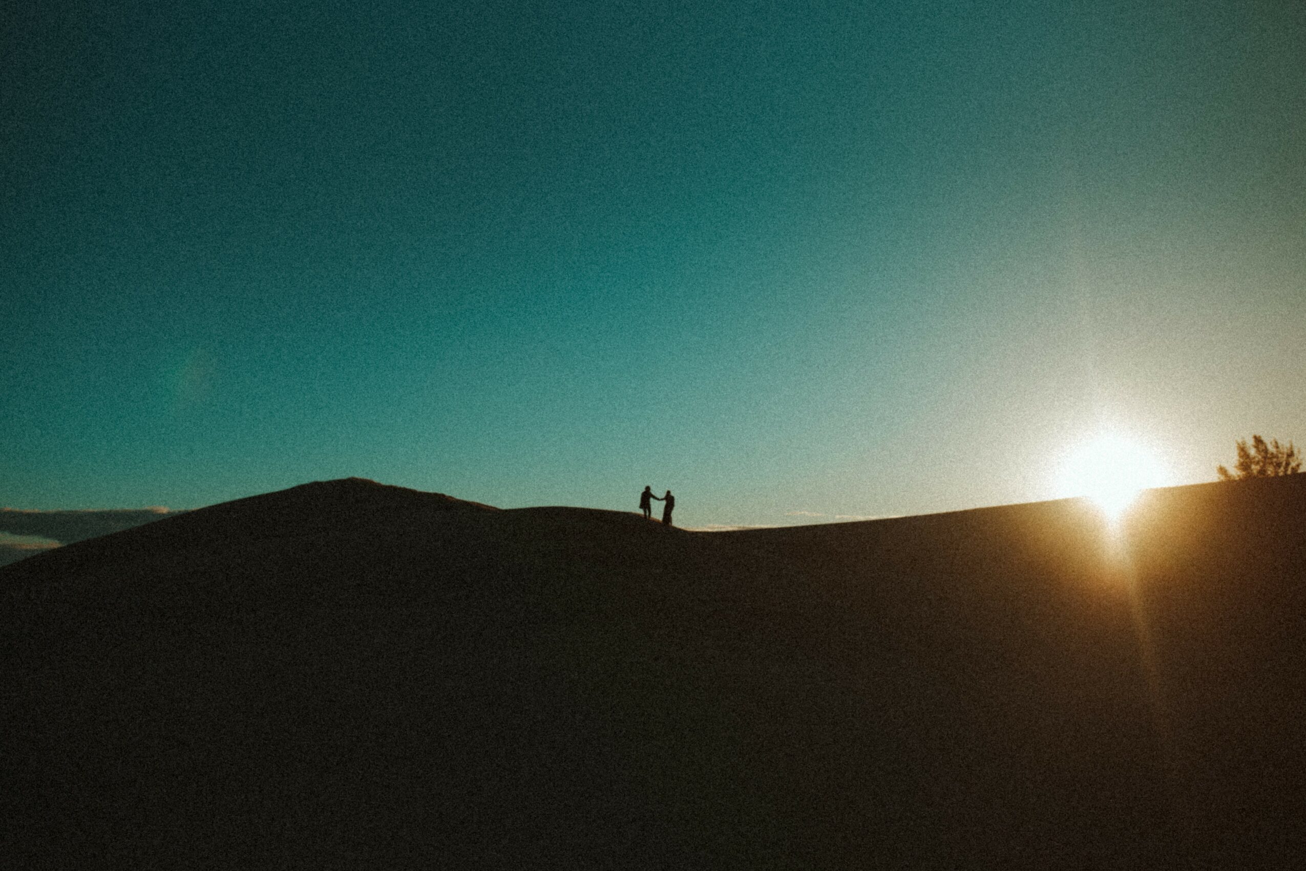 Silhouettes of two people standing on a hilltop against a bright sky with the sun near the horizon.