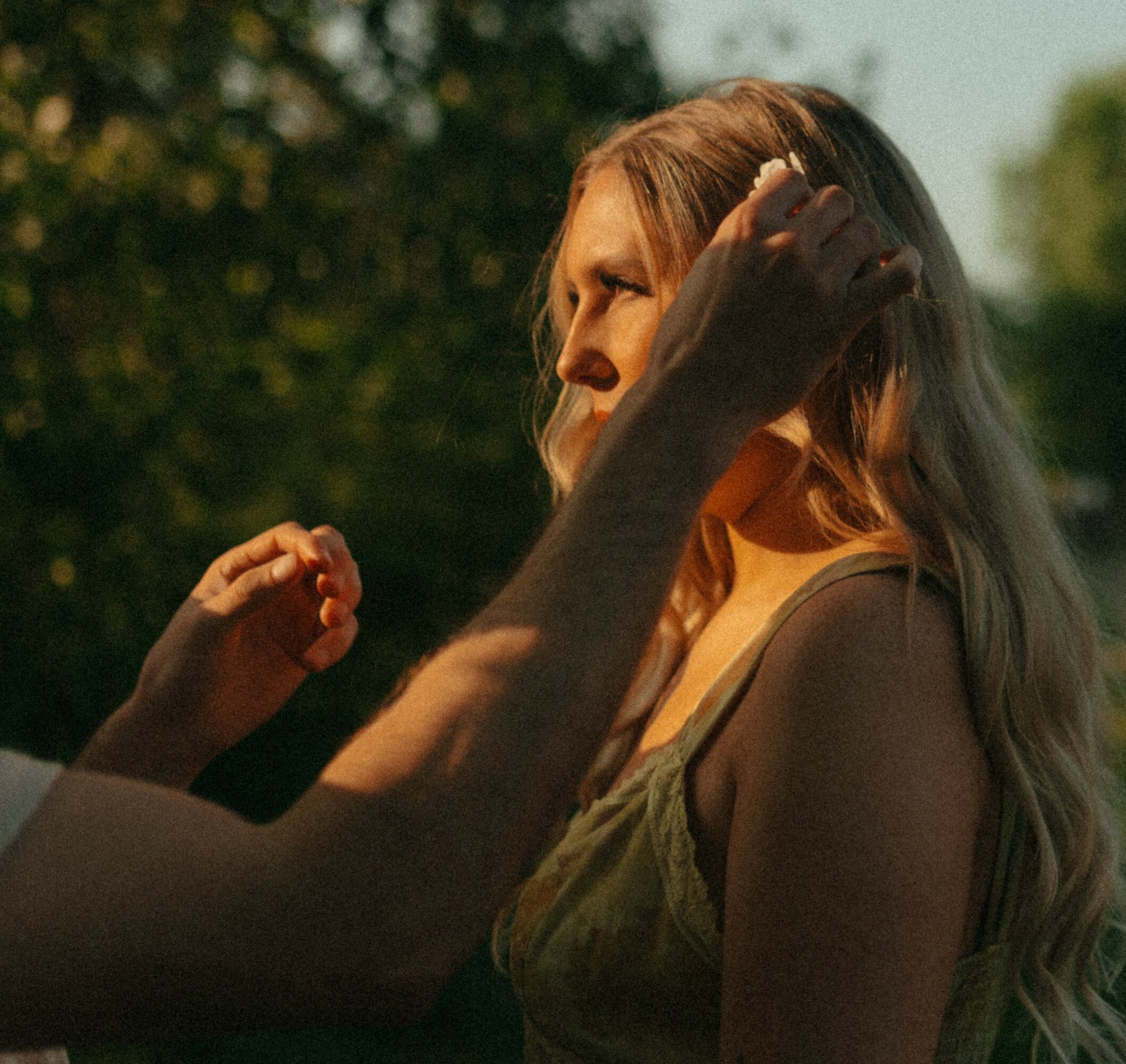 Person adjusting a woman's hair outdoors with trees in background, sunlight illuminating her face and hair.