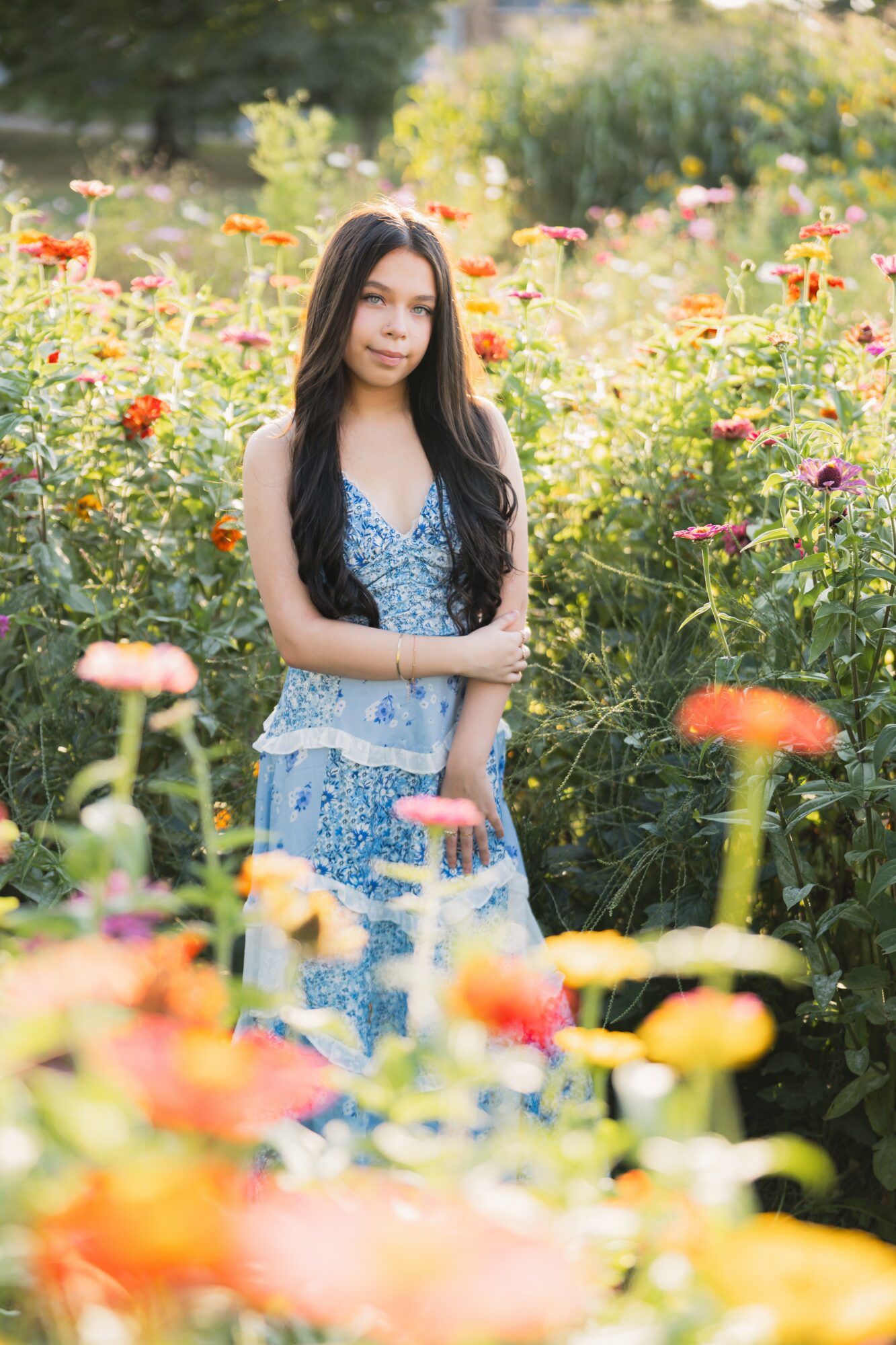 Young woman with long dark hair standing among colorful flowers in a garden, sunlight shining through trees.