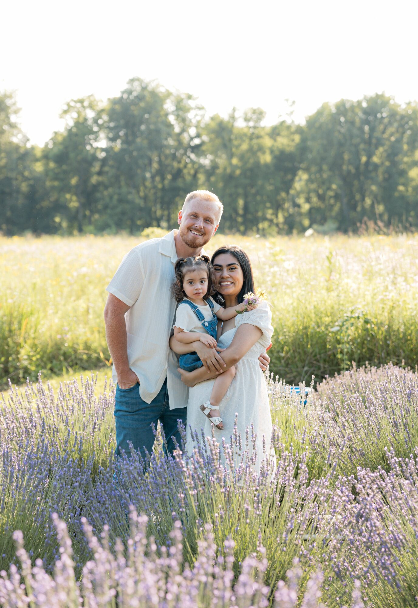 Family of three standing in a lavender field with trees in the background, smiling at the camera.