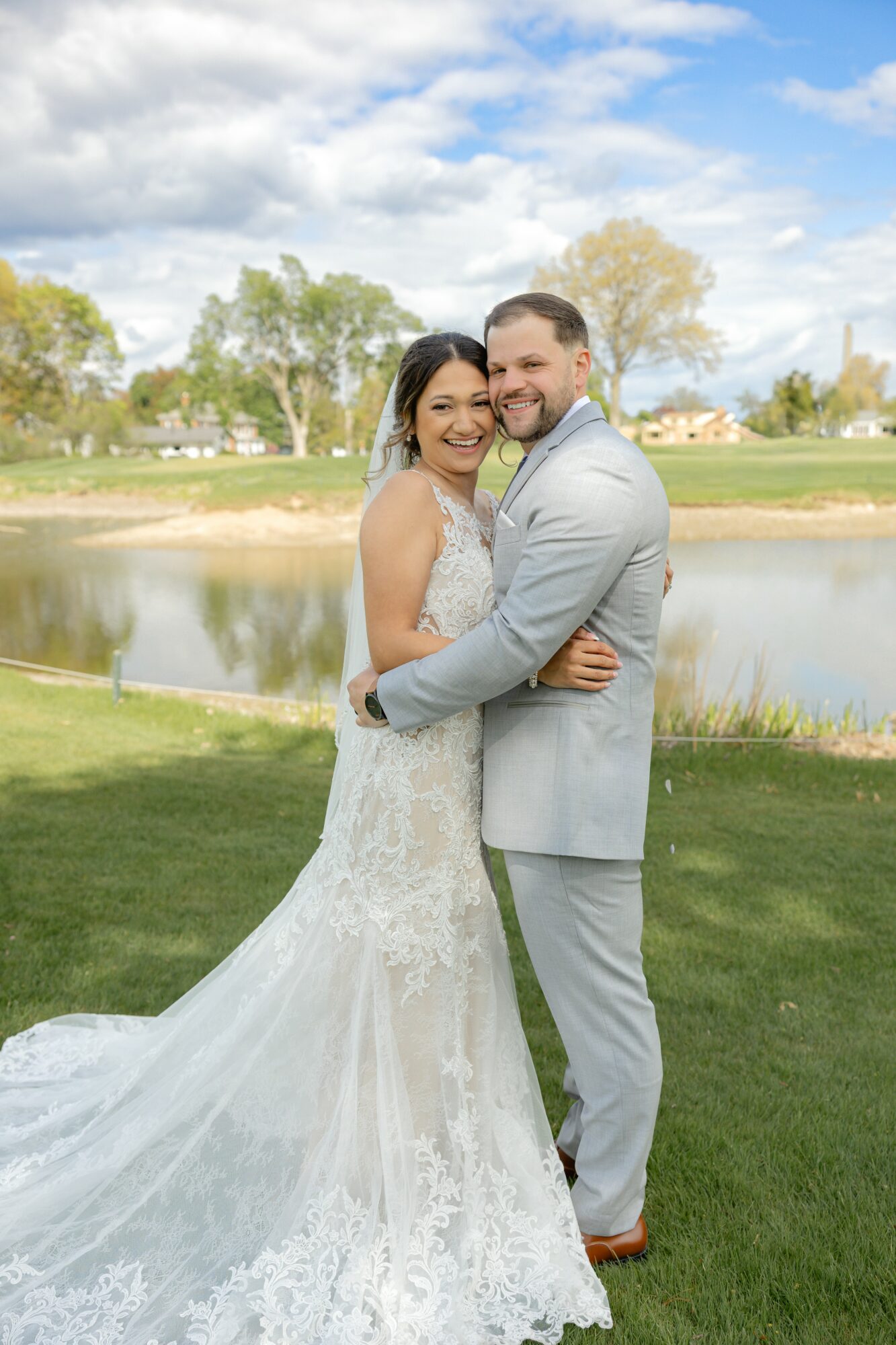 A bride and groom stand outdoors near a lake, smiling and embracing, with trees and a cloudy sky in the background.