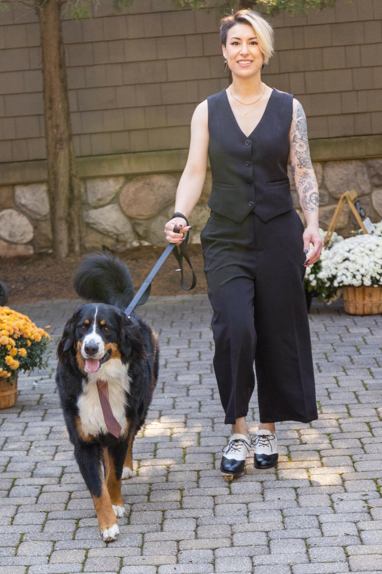 Woman walking a large dog outdoors on a paved path, with a wooden fence and flowers in the background.