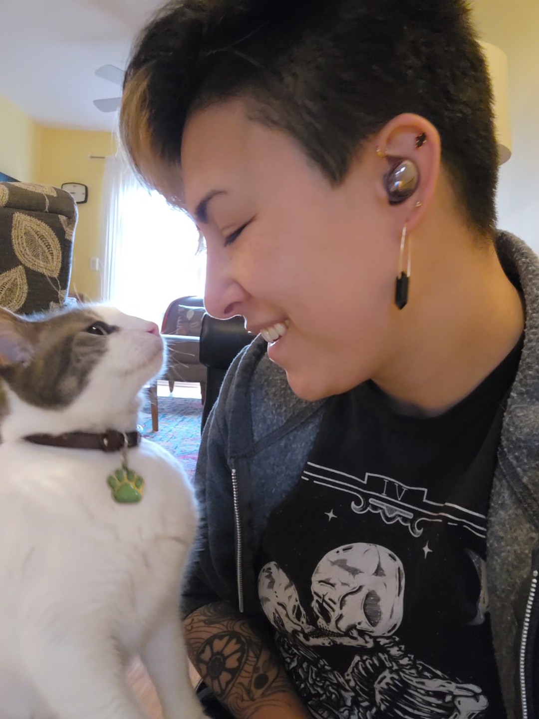 Person smiling with eyes closed, wearing earphones, close to a cat with a collar and tag, indoors with a window in the background.