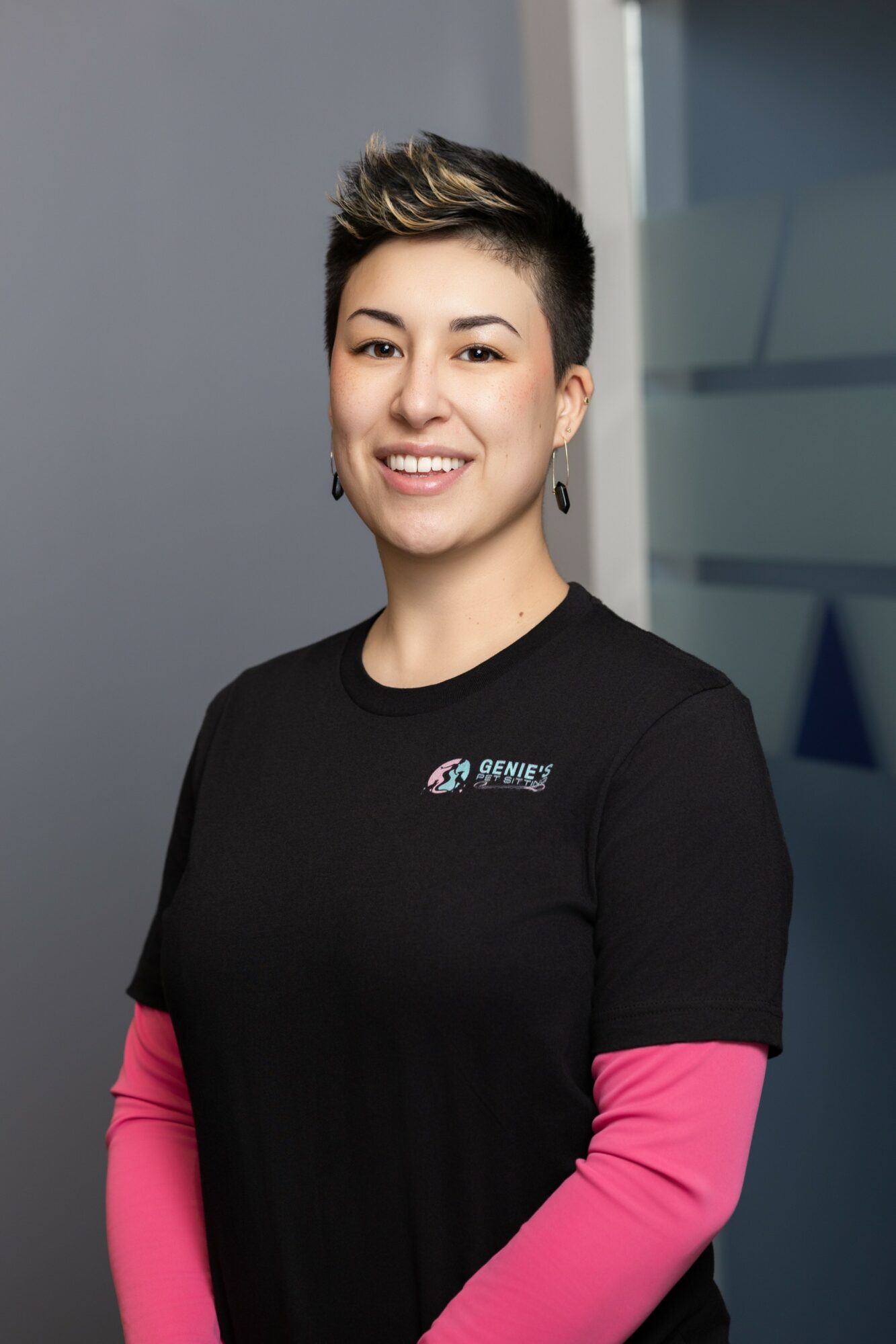Young woman with short hair smiling, wearing a black shirt with pink sleeves, standing indoors.