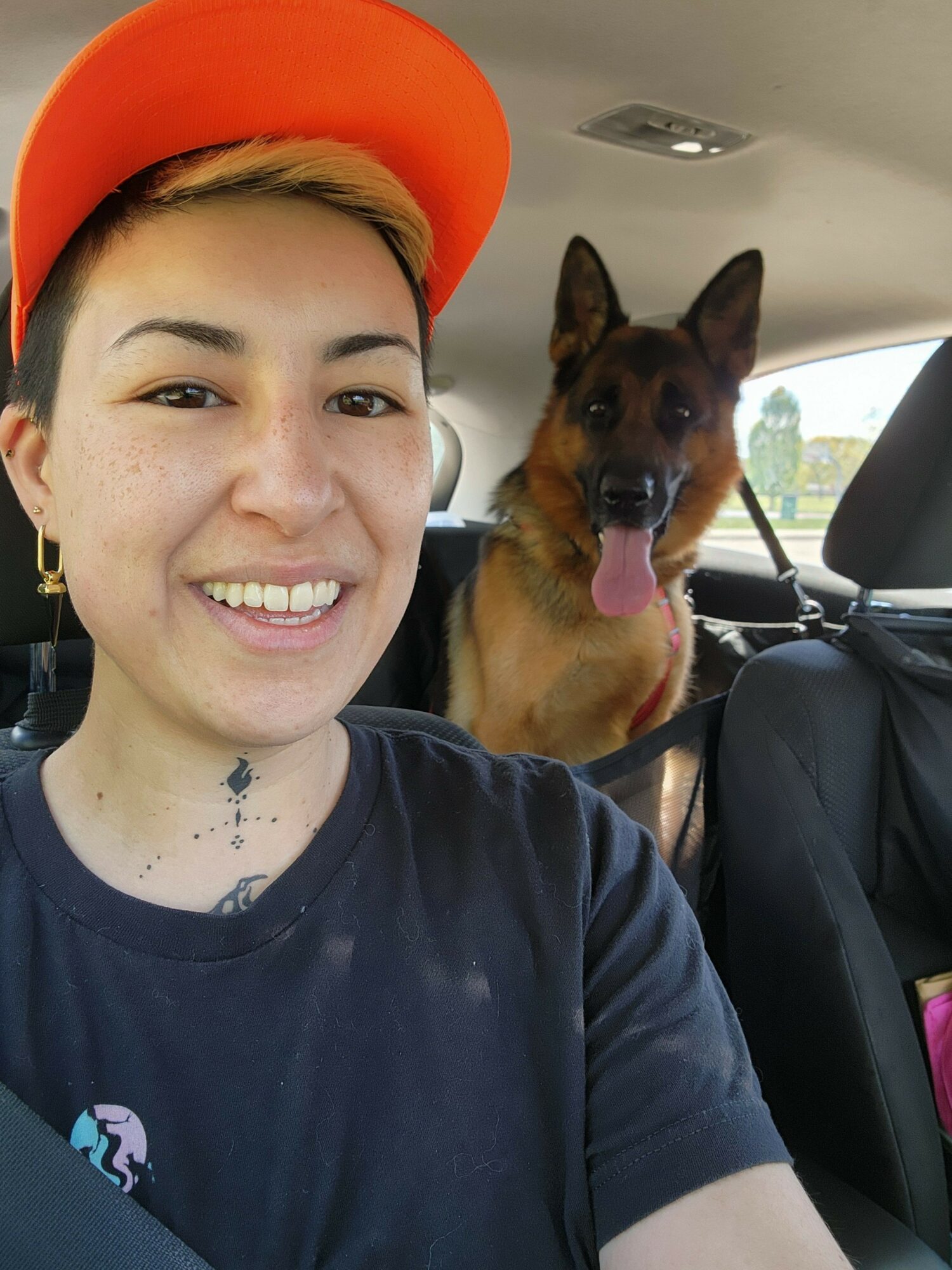 Person with short dark hair and orange cap smiling, dog with tongue out in car, seatbelt visible