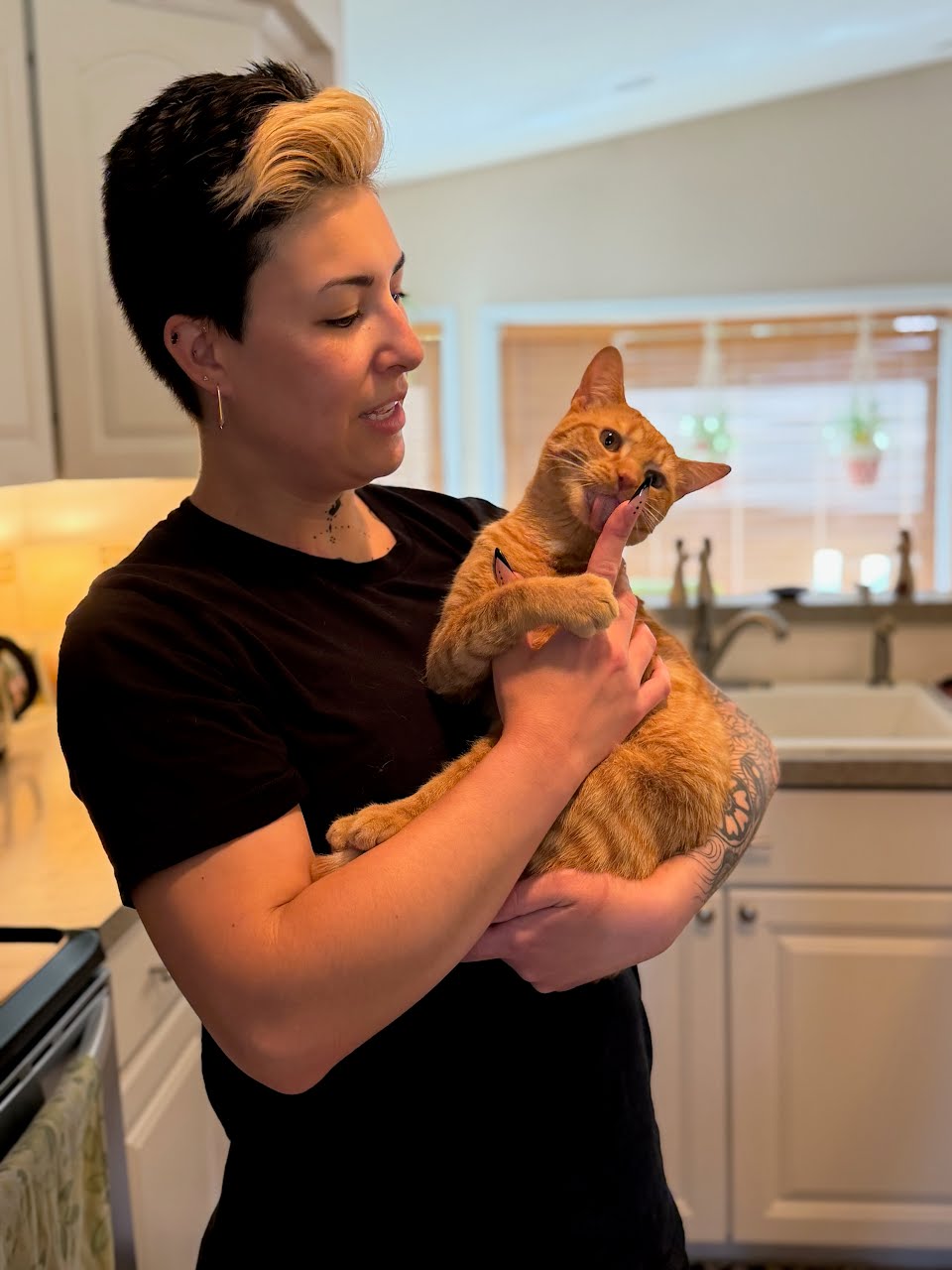 Woman holding an orange cat yawning in a kitchen with white cabinets and a window.