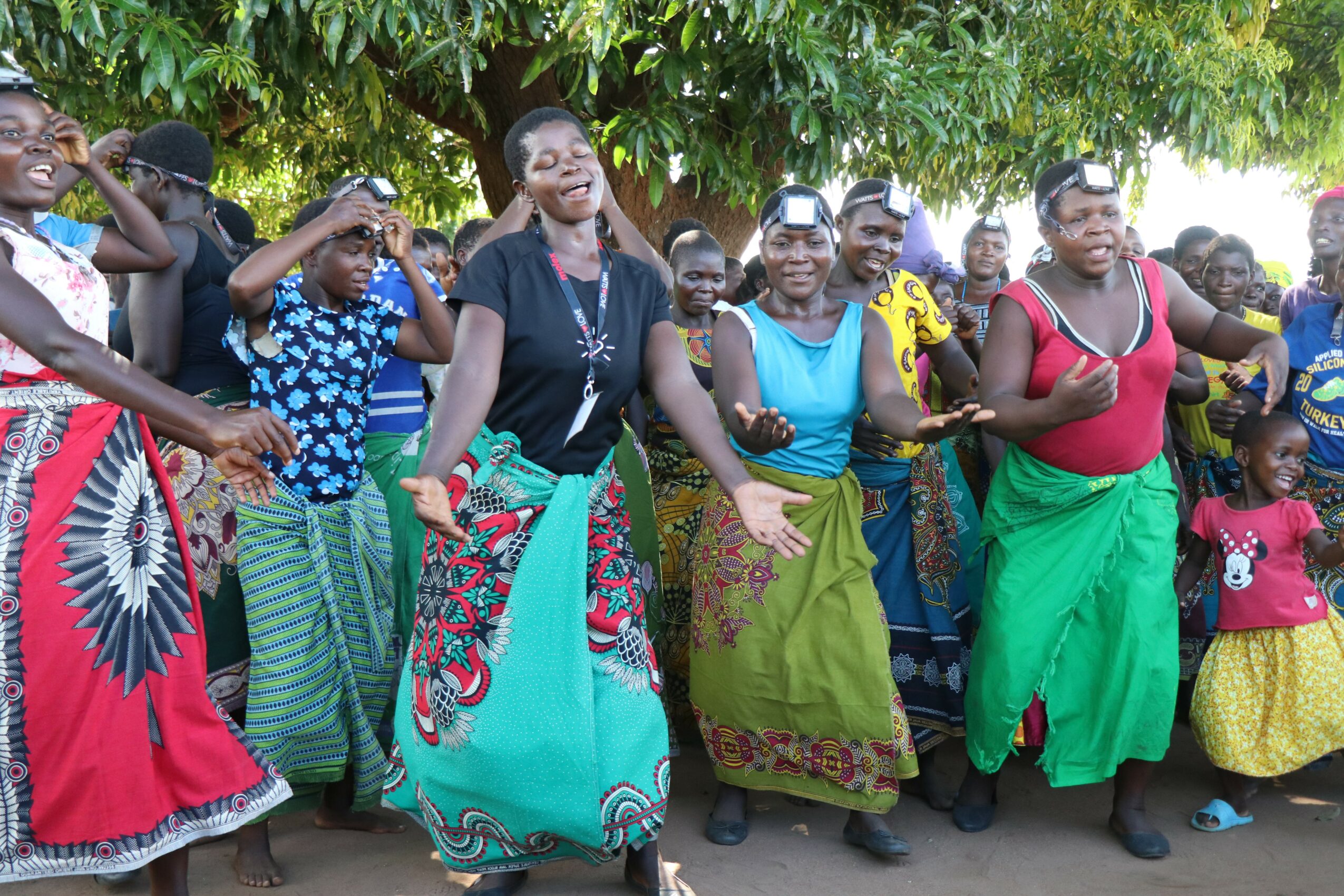 Group of women and children dancing outdoors under a tree, wearing colorful traditional clothing.