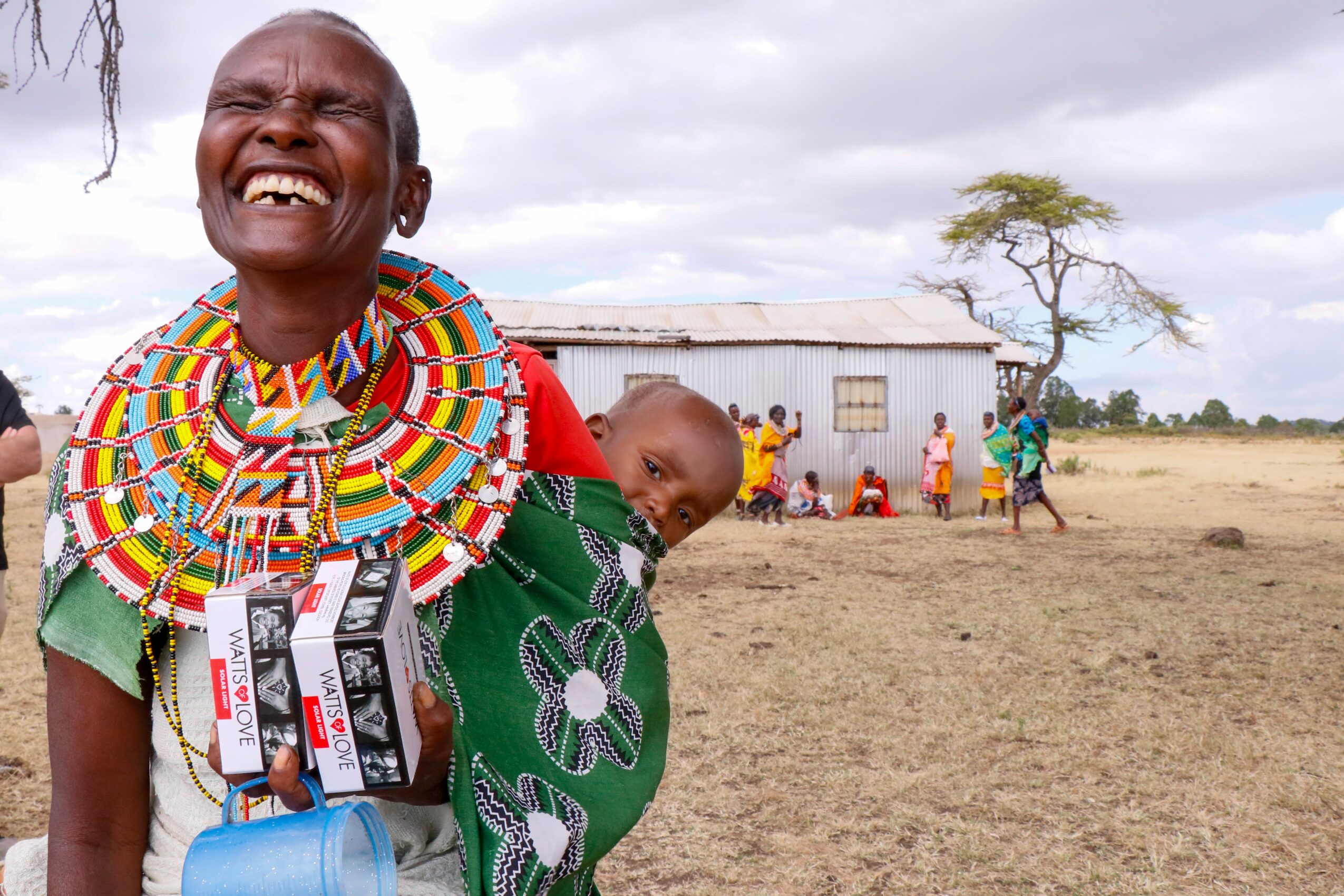 Smiling woman with colorful beaded jewelry holding a child on her back, in a rural setting with a house and people in the background.