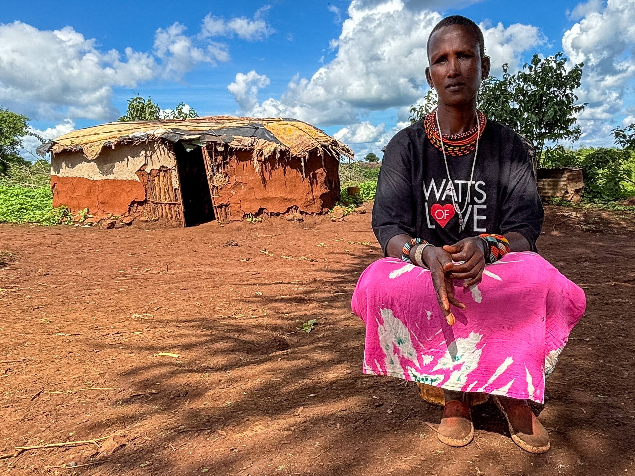 Woman sitting outdoors on reddish ground with a small mud house and trees in background, under a partly cloudy sky.