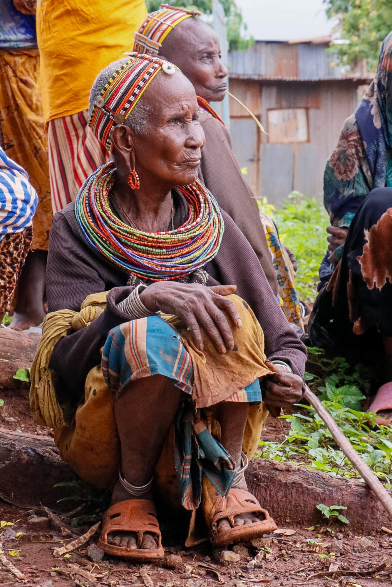 Elderly woman with traditional jewelry and clothing, sitting outdoors with other people nearby.