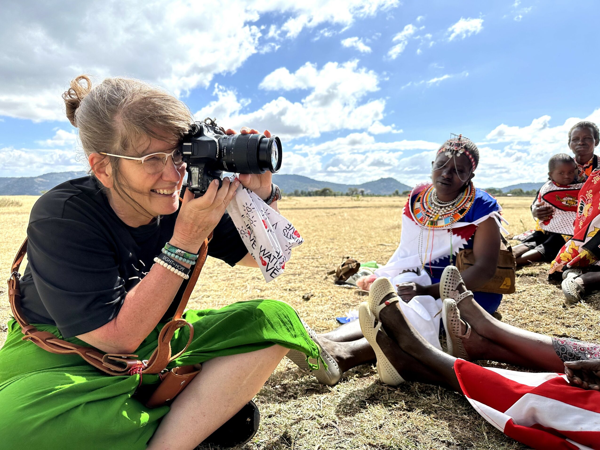 Woman with glasses taking a photo outdoors, sitting on the ground with a group of people in traditional clothing nearby, under a blue sky.