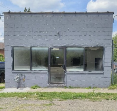Gray building with large front windows and a door, situated on a grassy area under a partly cloudy sky.