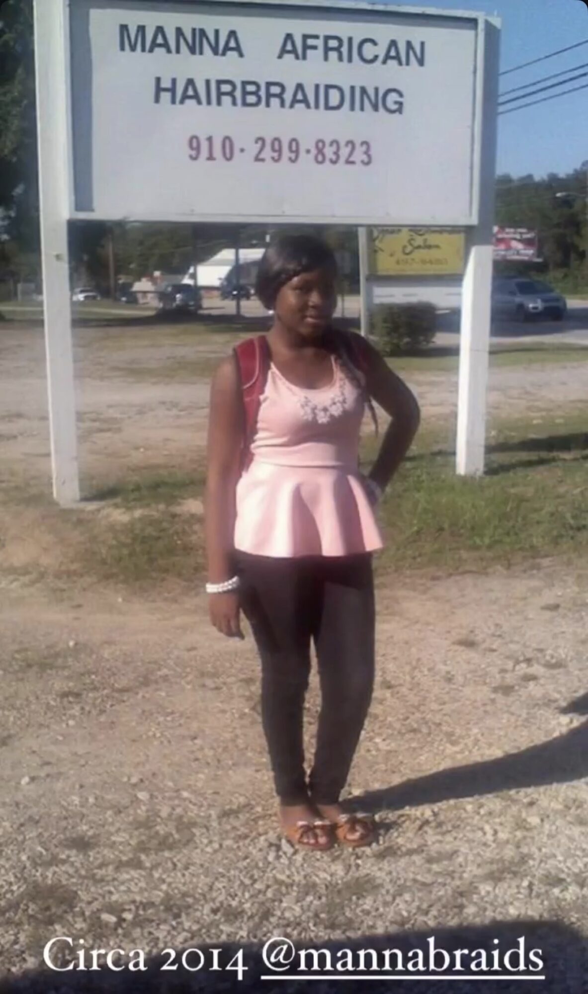 Young girl standing outdoors in front of a sign for a hairbraiding business, wearing a pink top and black pants.