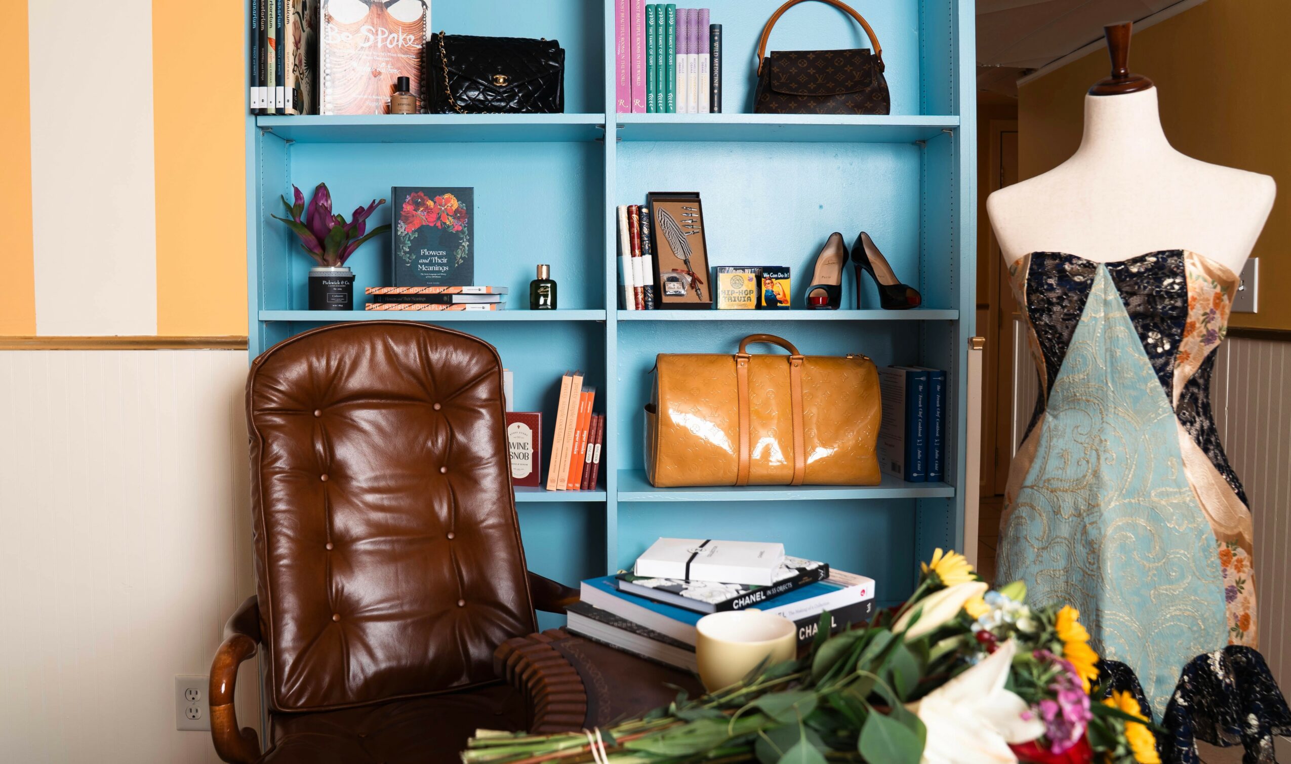 Brown leather chair in front of a blue bookshelf filled with books and decorative items, next to a mannequin with fabric and a dress.