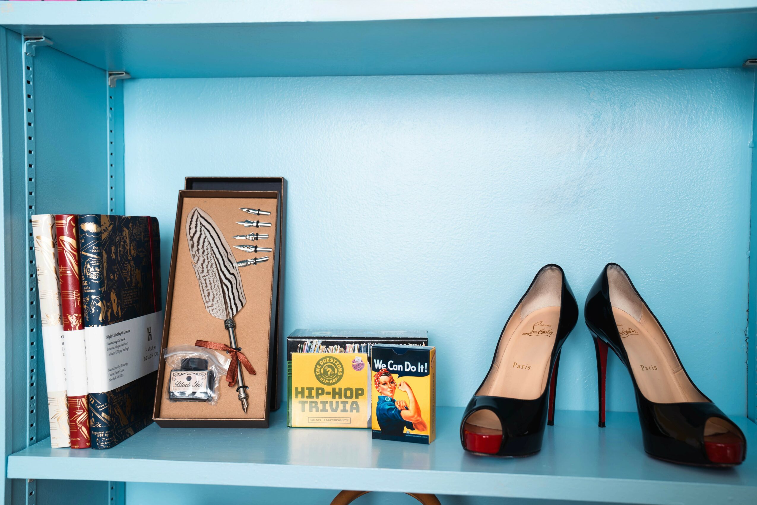Shelf with books, framed picture, a small box, and a pair of black high heels with red soles, against a blue wall.
