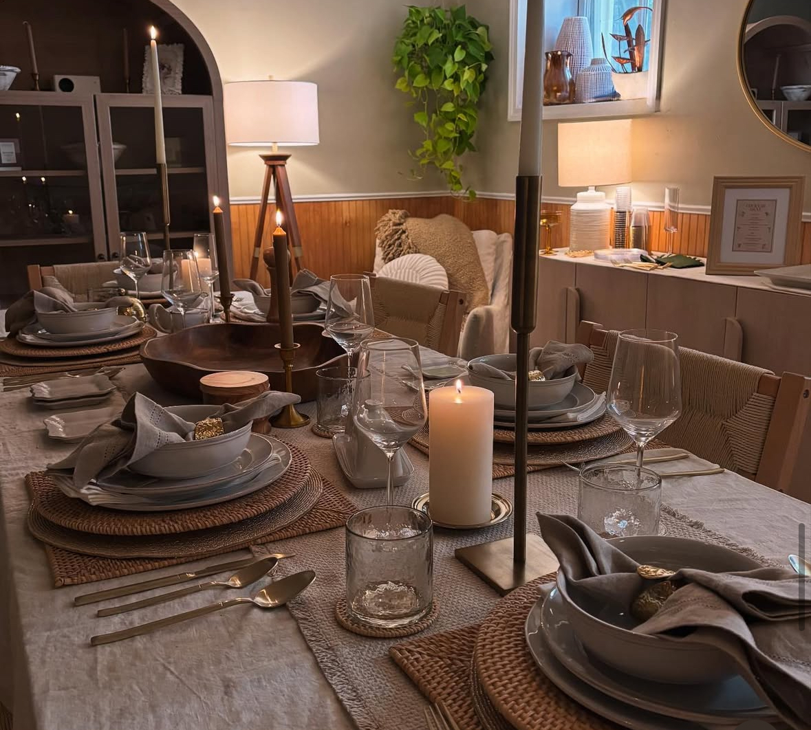 Dining table set with plates, glasses, cutlery, and candles in a cozy, warmly lit room with a window and decorative items.