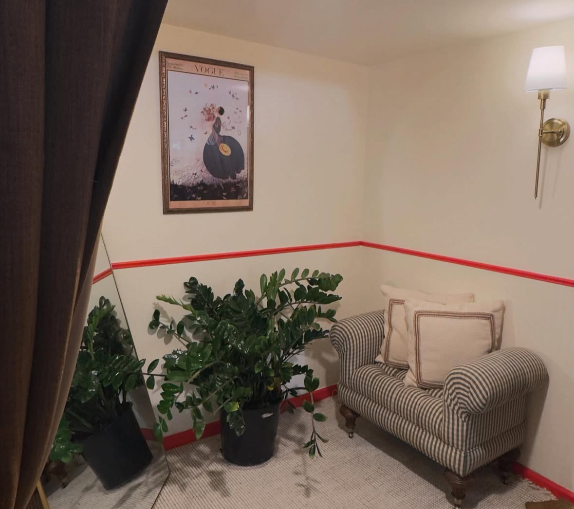 Corner of a room with a striped armchair, potted plant, framed artwork, and wall-mounted lamp, with a red stripe on the wall.