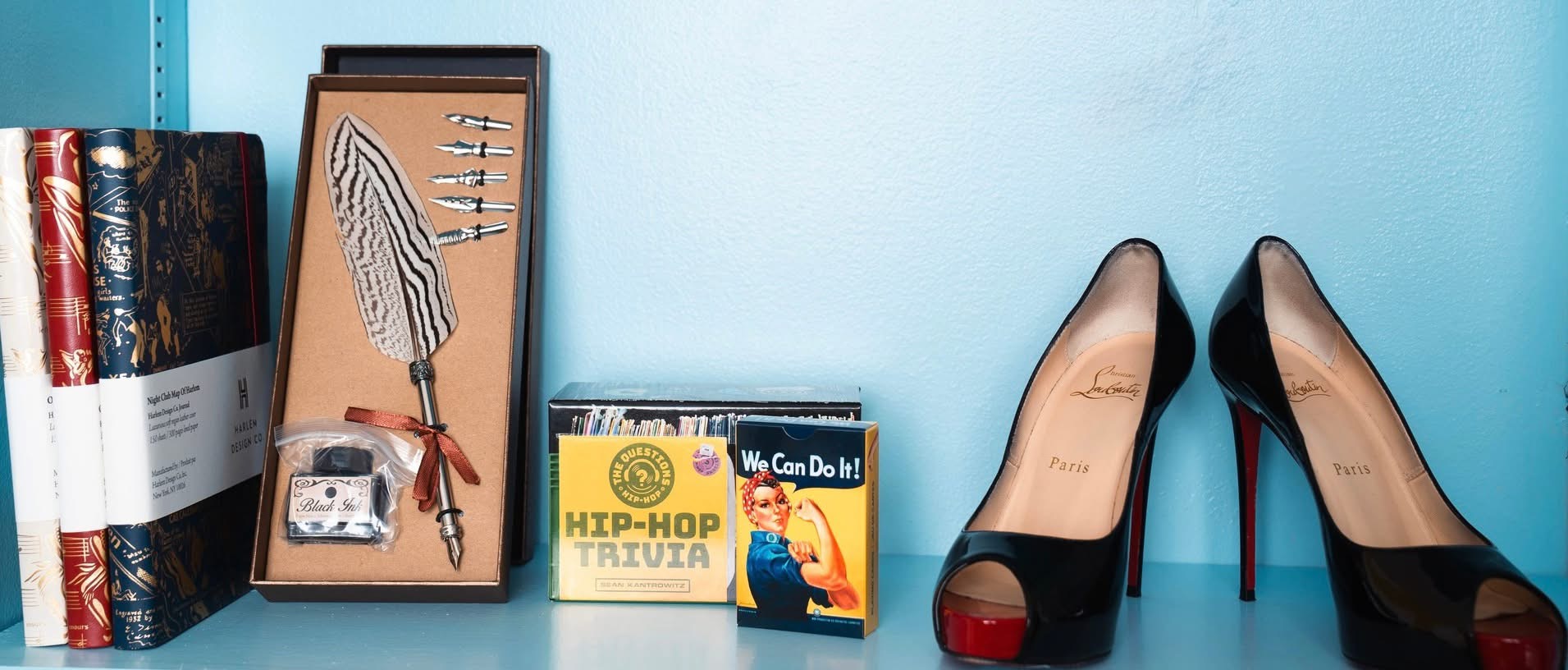 Pair of black high heels with red soles, books, framed artwork, and a small box on a blue surface.
