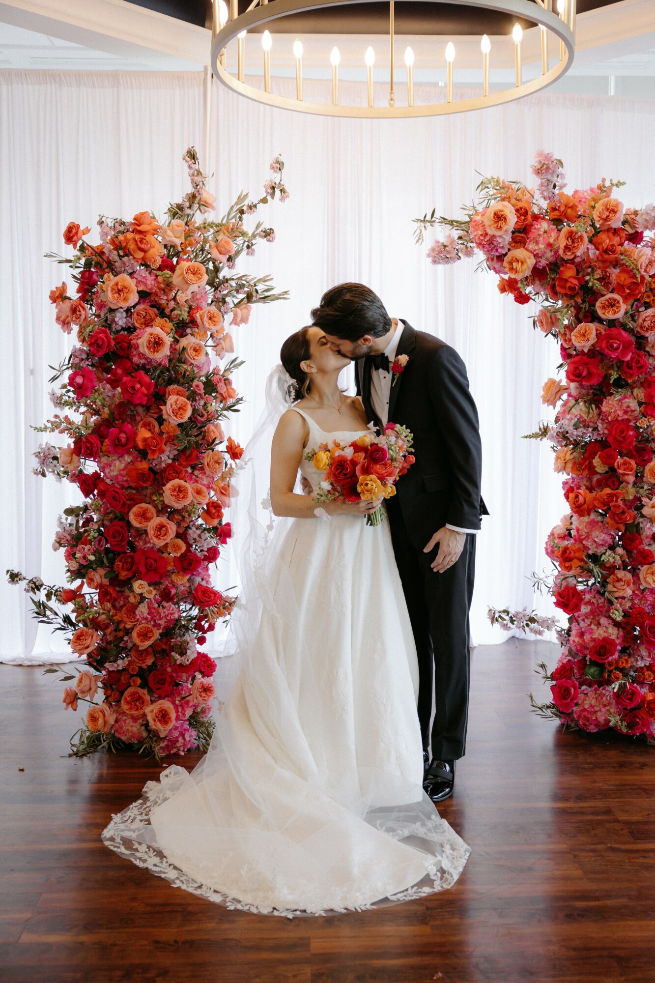 Bride and groom stand close, holding a bouquet, surrounded by large floral arrangements, in a bright room with white curtains.