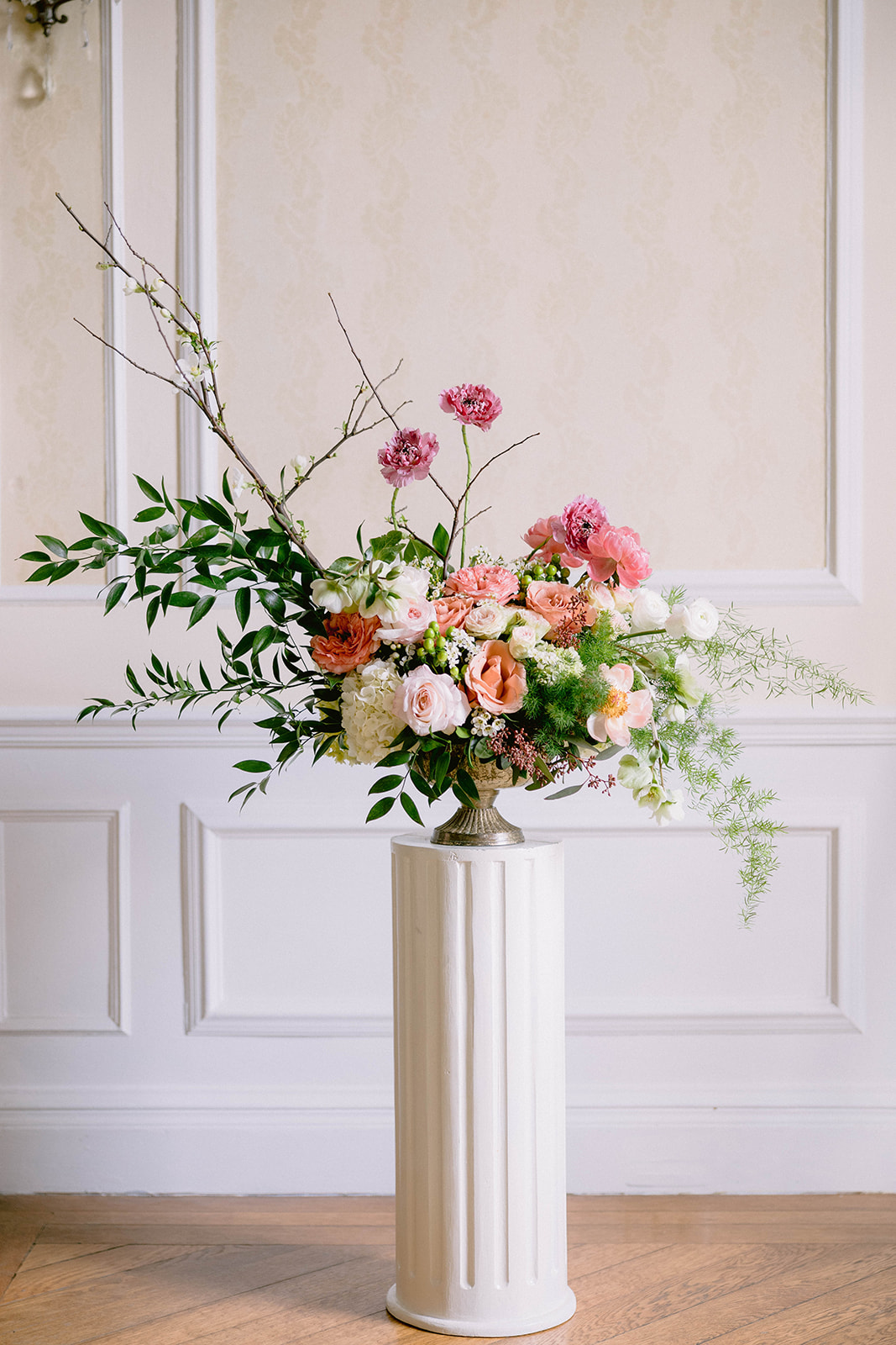 Flower arrangement with pink, white, and peach blooms in a vase on a white pedestal against a beige wall.