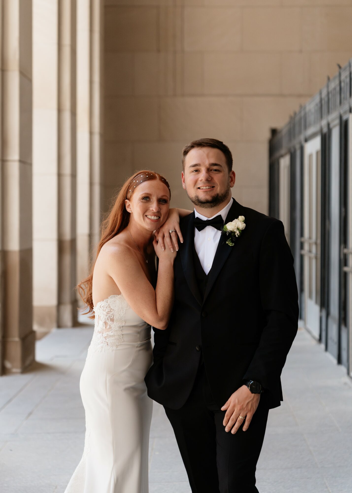 A smiling couple dressed in wedding attire standing outdoors near a building with columns and glass windows.