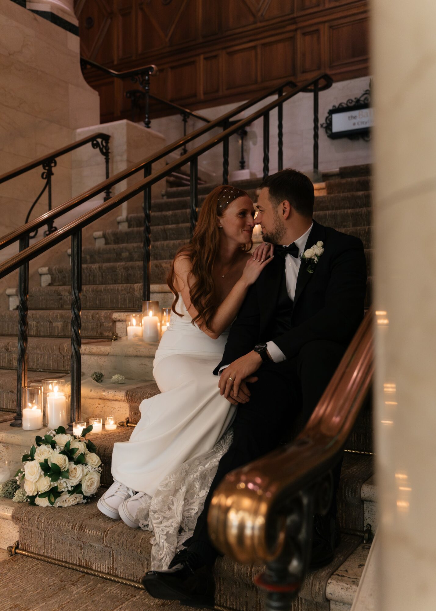 Bride and groom sitting on stairs, close together, in a wedding setting with candles and flowers nearby.