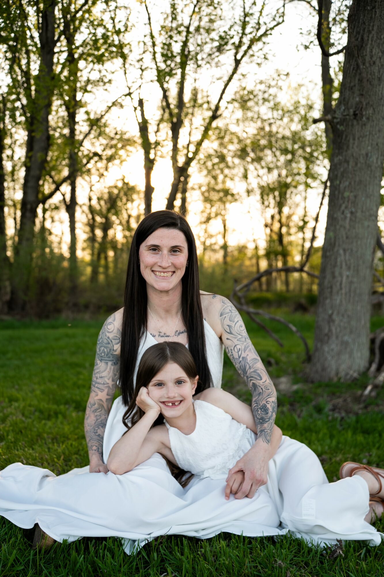 Woman and girl sitting on grass outdoors, smiling, trees in background, sunlight filtering through, woman wearing white dress, girl resting head on woman's lap.
